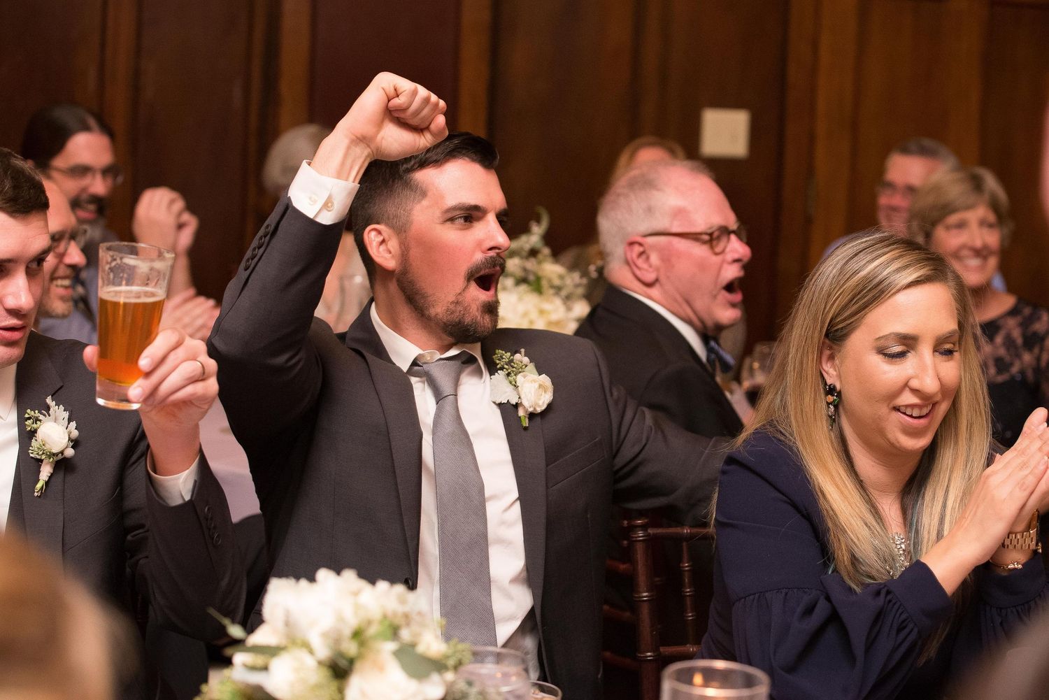 Excited groomsman raises fist and beer glass during lively wedding reception celebration at elegant venue.