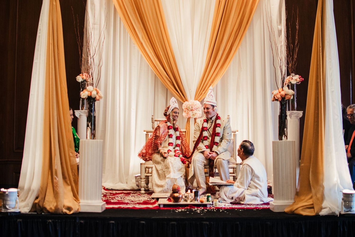 Indian wedding ceremony with couple seated under ornate gold and white mandap with floral decorations.