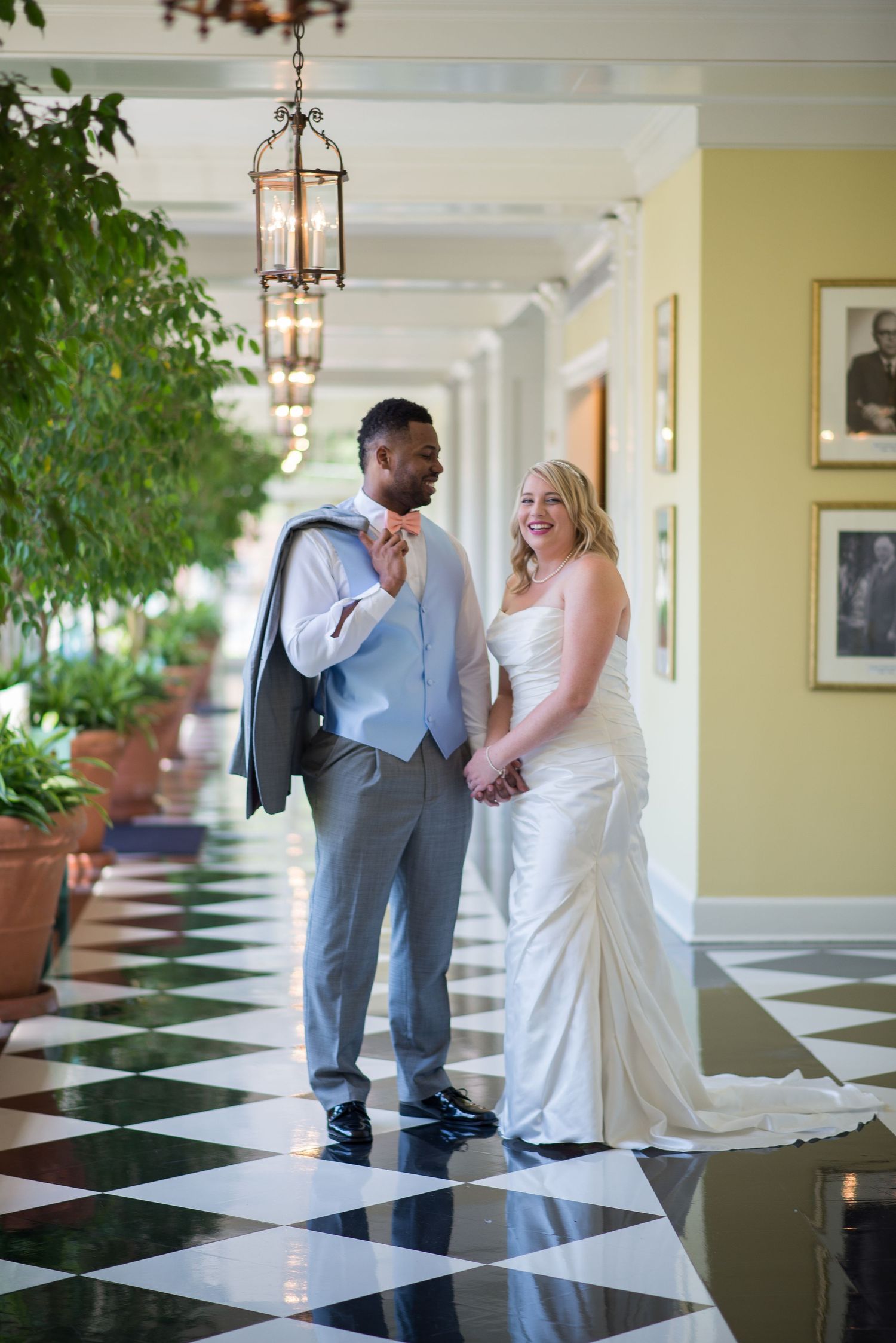 Interracial newlyweds hold hands and smile in elegant hallway with black and white checkered floor.