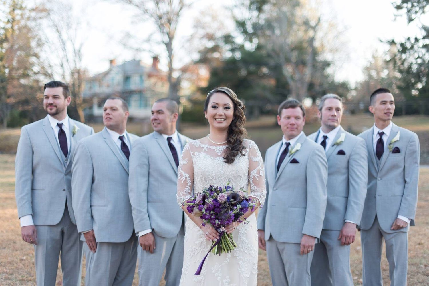 Bride holding purple bouquet stands before groomsmen in gray suits outdoors.