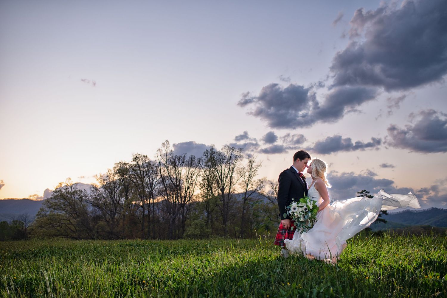 Bride and groom kiss at sunset in scenic mountain meadow.