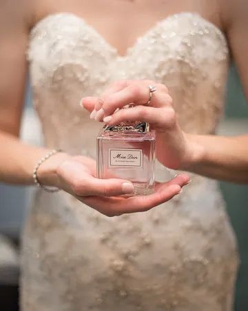 Bride in lace gown holds Miss Dior perfume bottle with pearl bracelet.