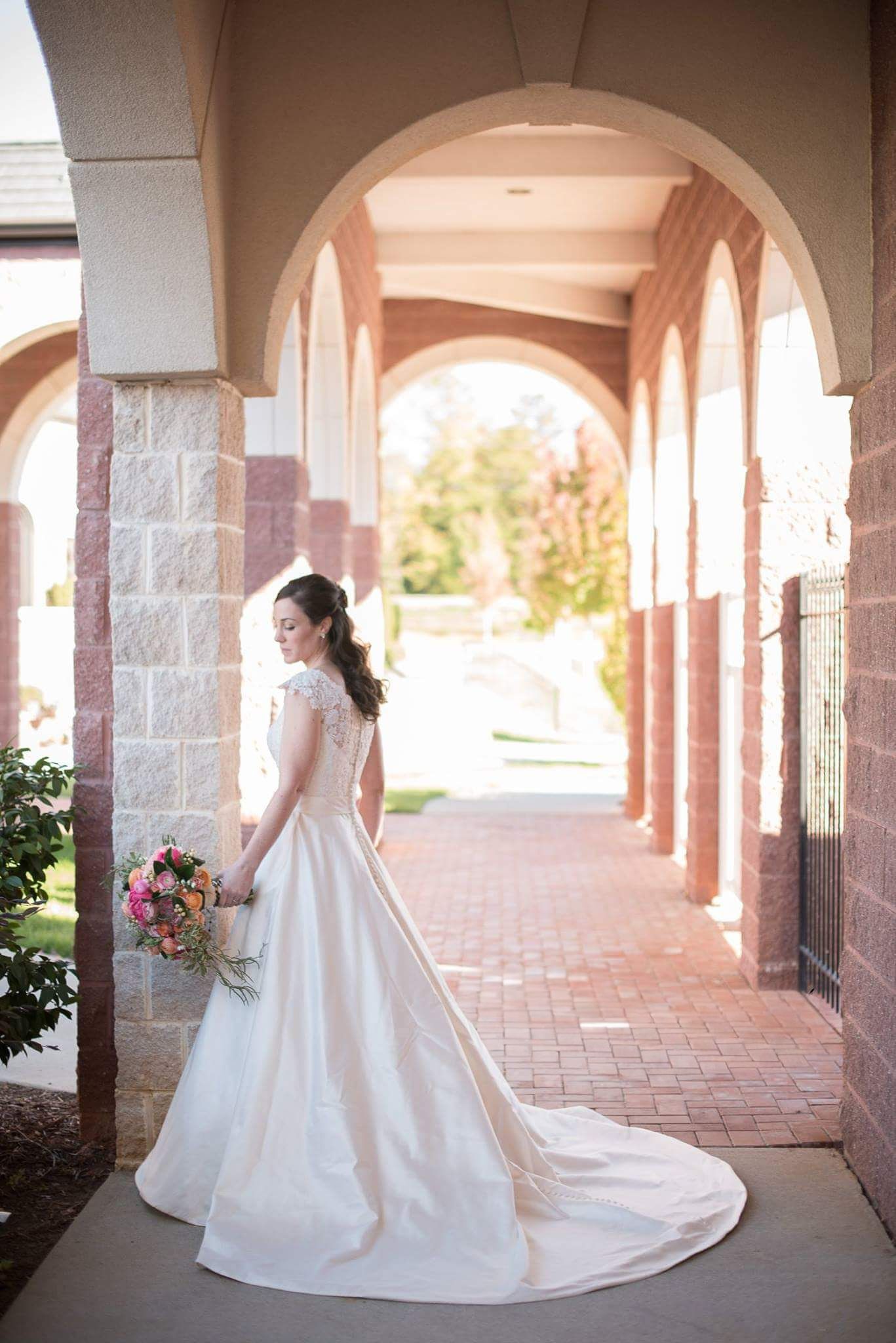 An elegant bride in a lace gown poses under arched columns holding a colorful bouquet at a church venue.