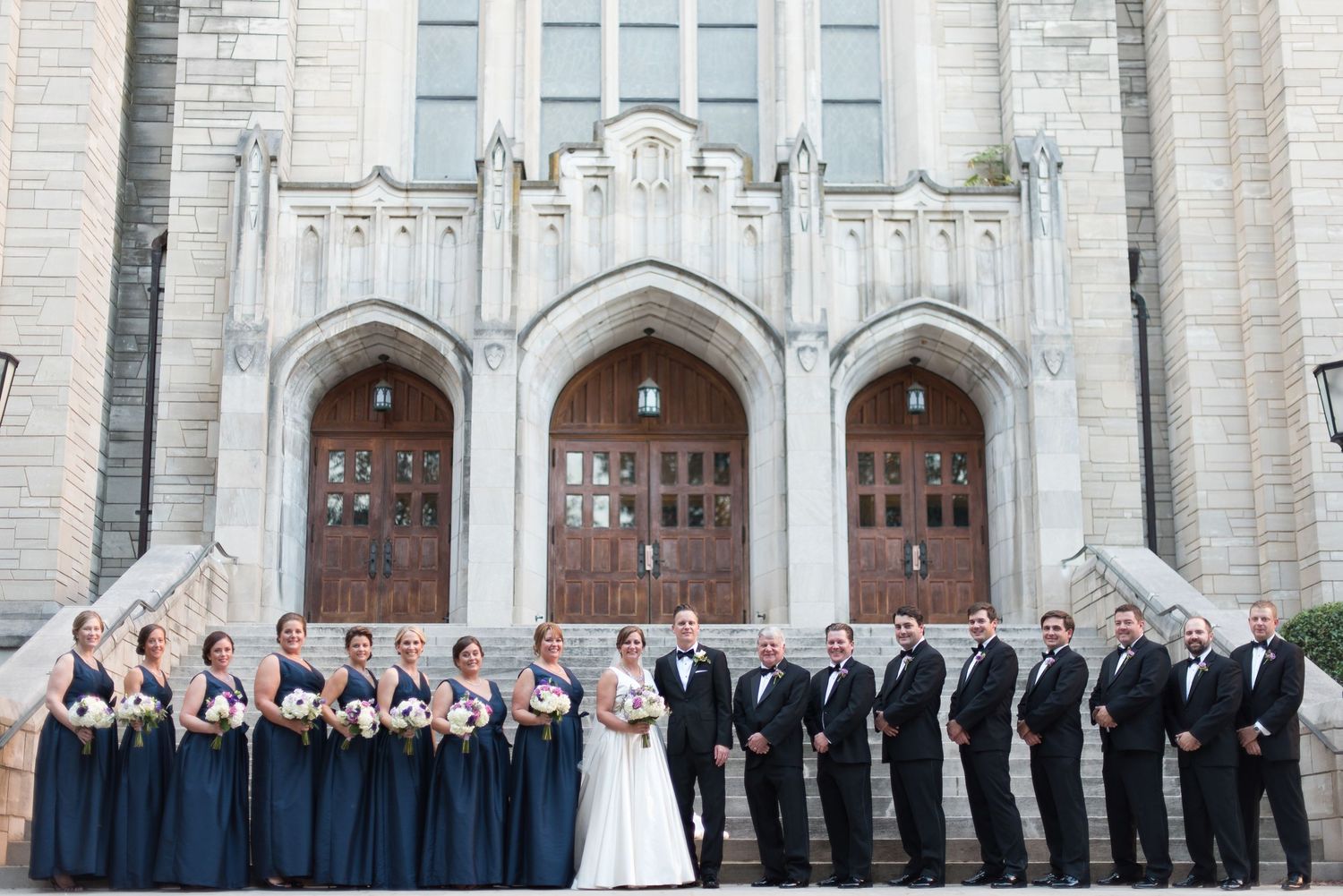 Wedding party in navy and black formal attire posing before gothic stone church with arched wooden doors.
