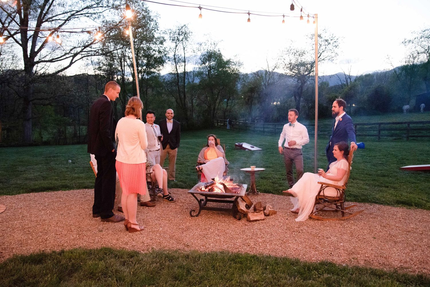 Guests gather around an outdoor fire pit at a rustic evening wedding reception with string lights and green hills.