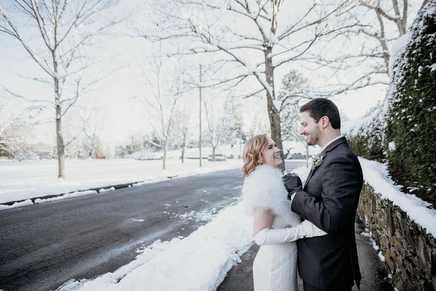 A bride and groom embrace in a snowy winter landscape, sharing a romantic moment beside a snow-covered stone wall.