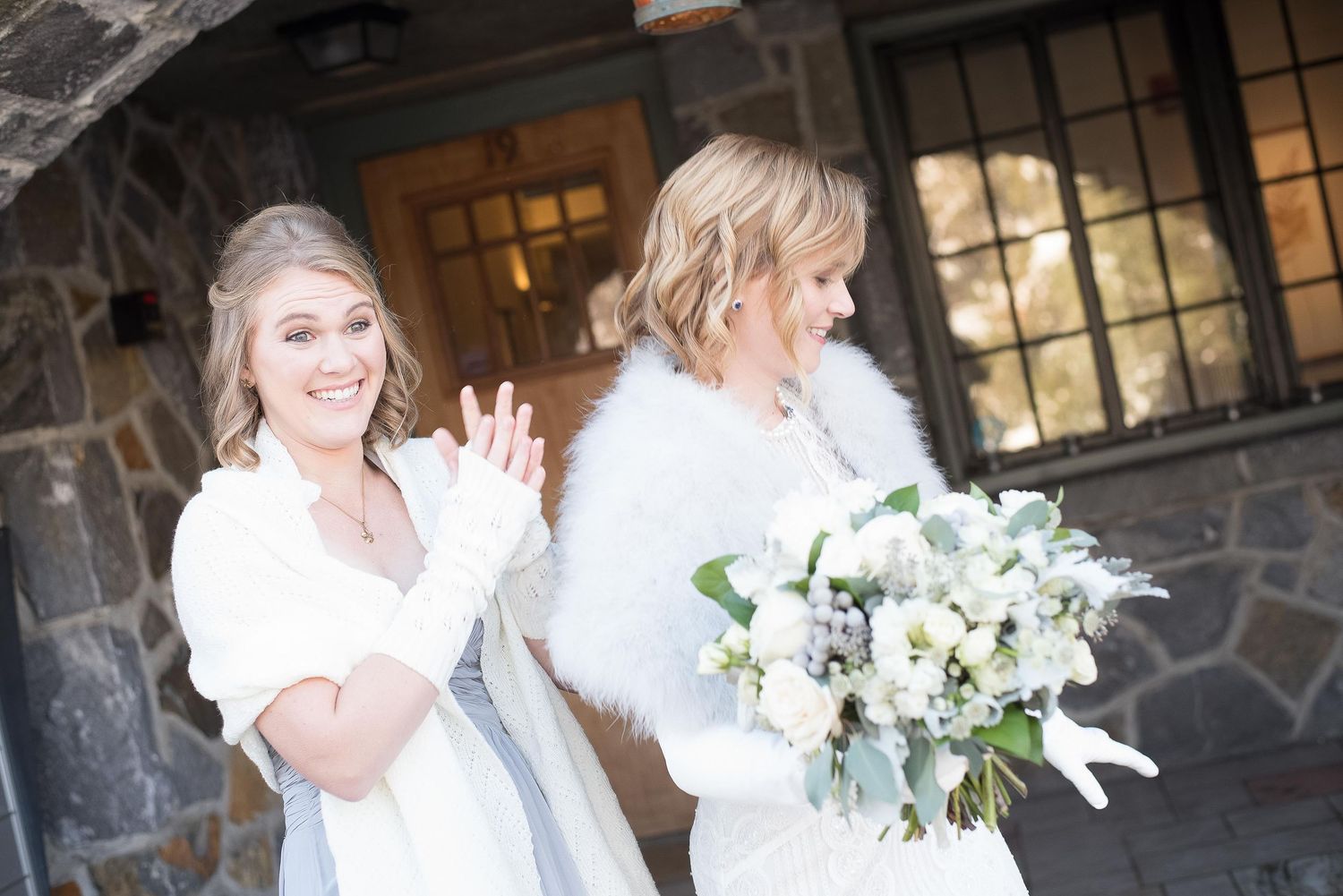 Two women in white winter wraps share a joyful moment outdoors at a stone wedding venue.
