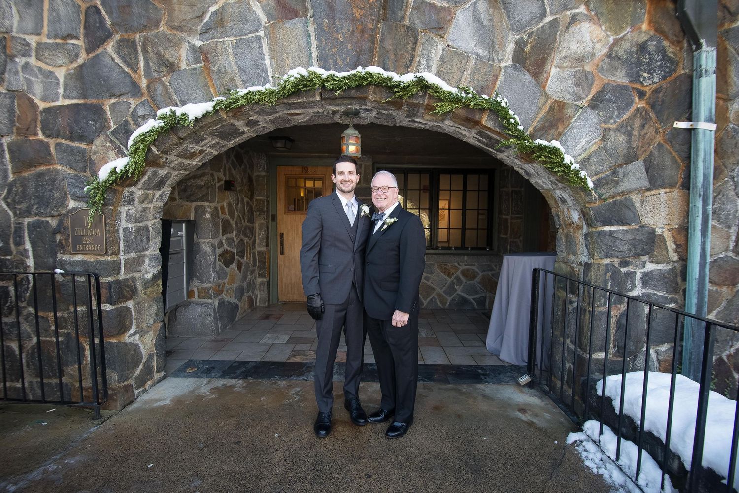 Two grooms pose under a stone archway at their winter wedding venue with snow on the ground.
