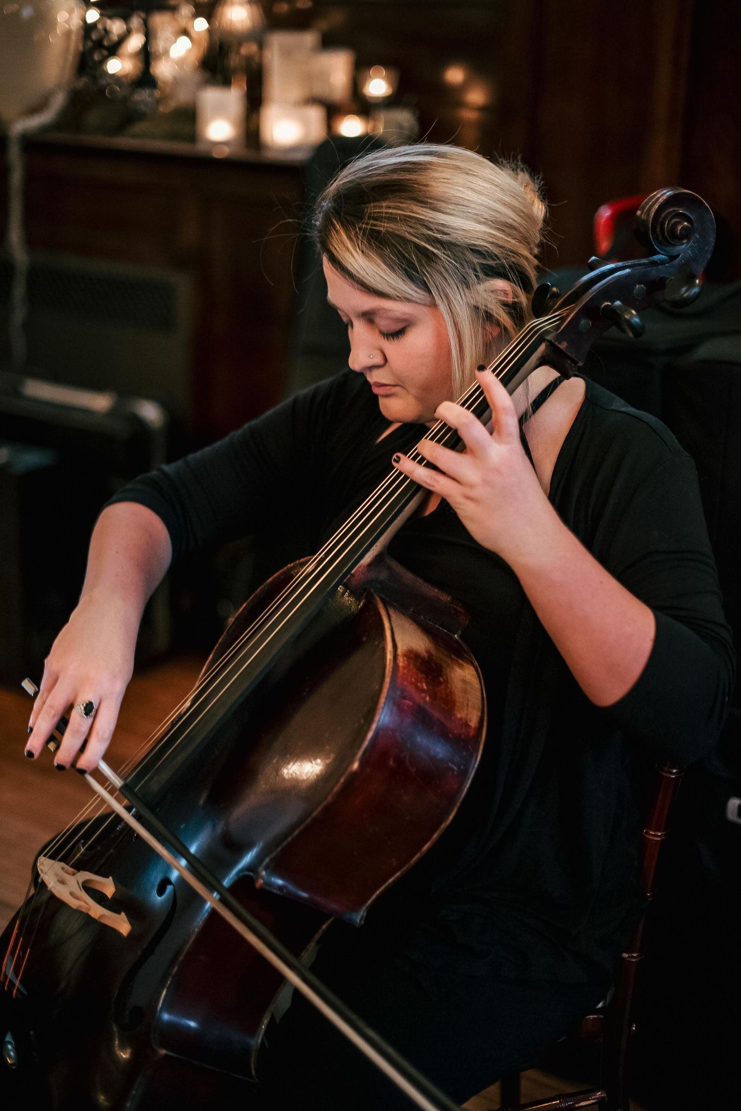 Young woman playing cello in dimly lit room with candles in background, focused on her performance.