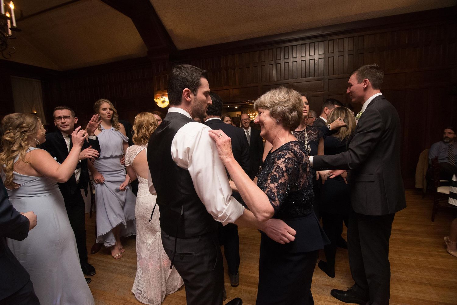 Guests dancing at a formal wedding reception in an elegant wood-paneled ballroom, wearing suits and gowns.
