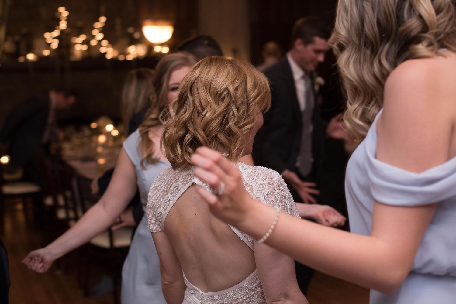 Bride in backless lace wedding dress dancing with guests at elegant wedding reception.