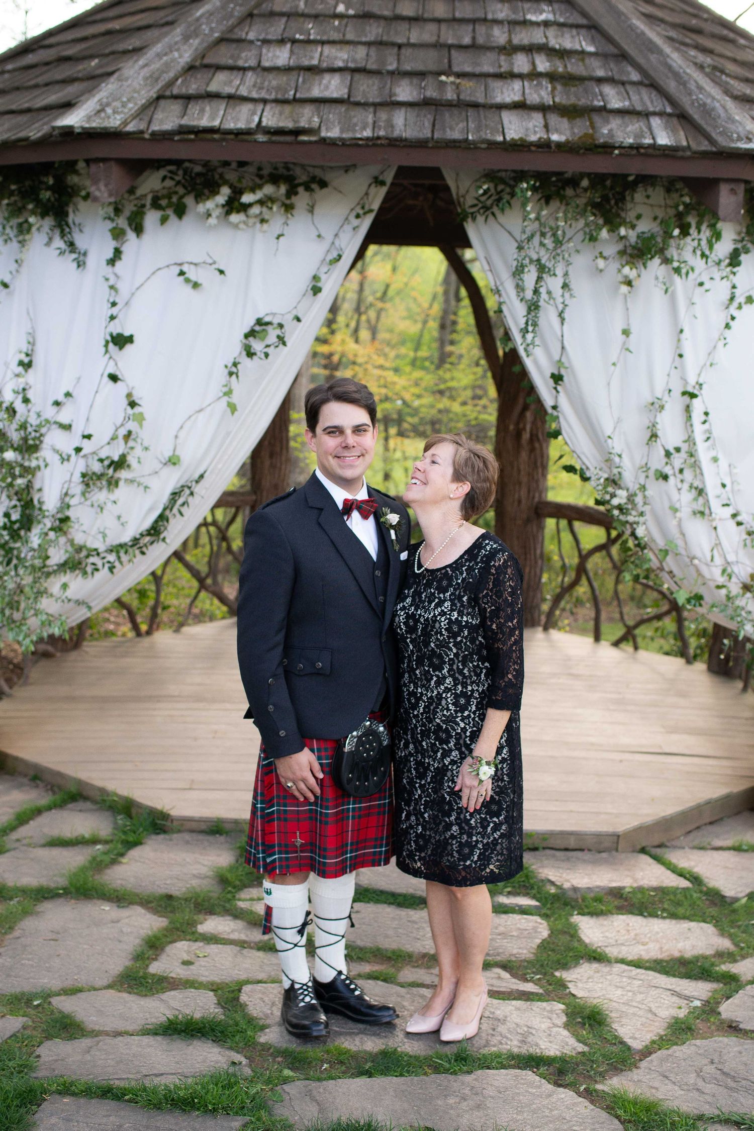 Groom in Scottish tartan kilt and woman in black lace dress pose under a vine-covered gazebo at an outdoor wedding.