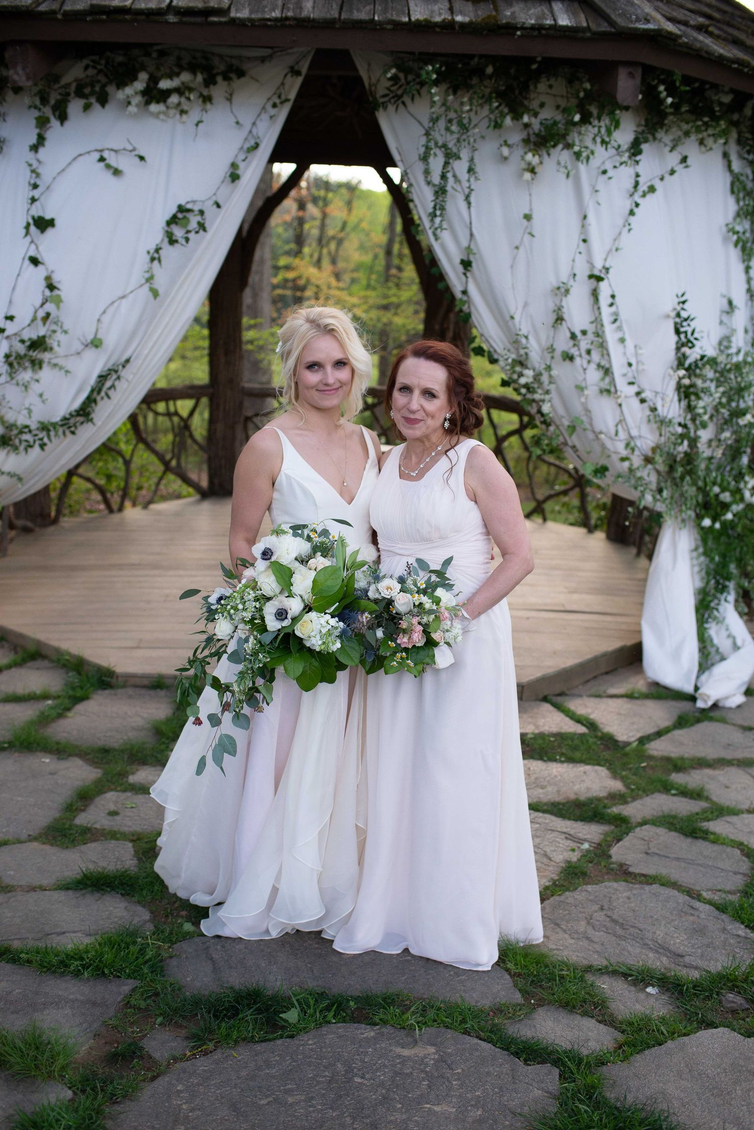 Two brides in white gowns holding bouquets stand before a floral gazebo on a stone path.