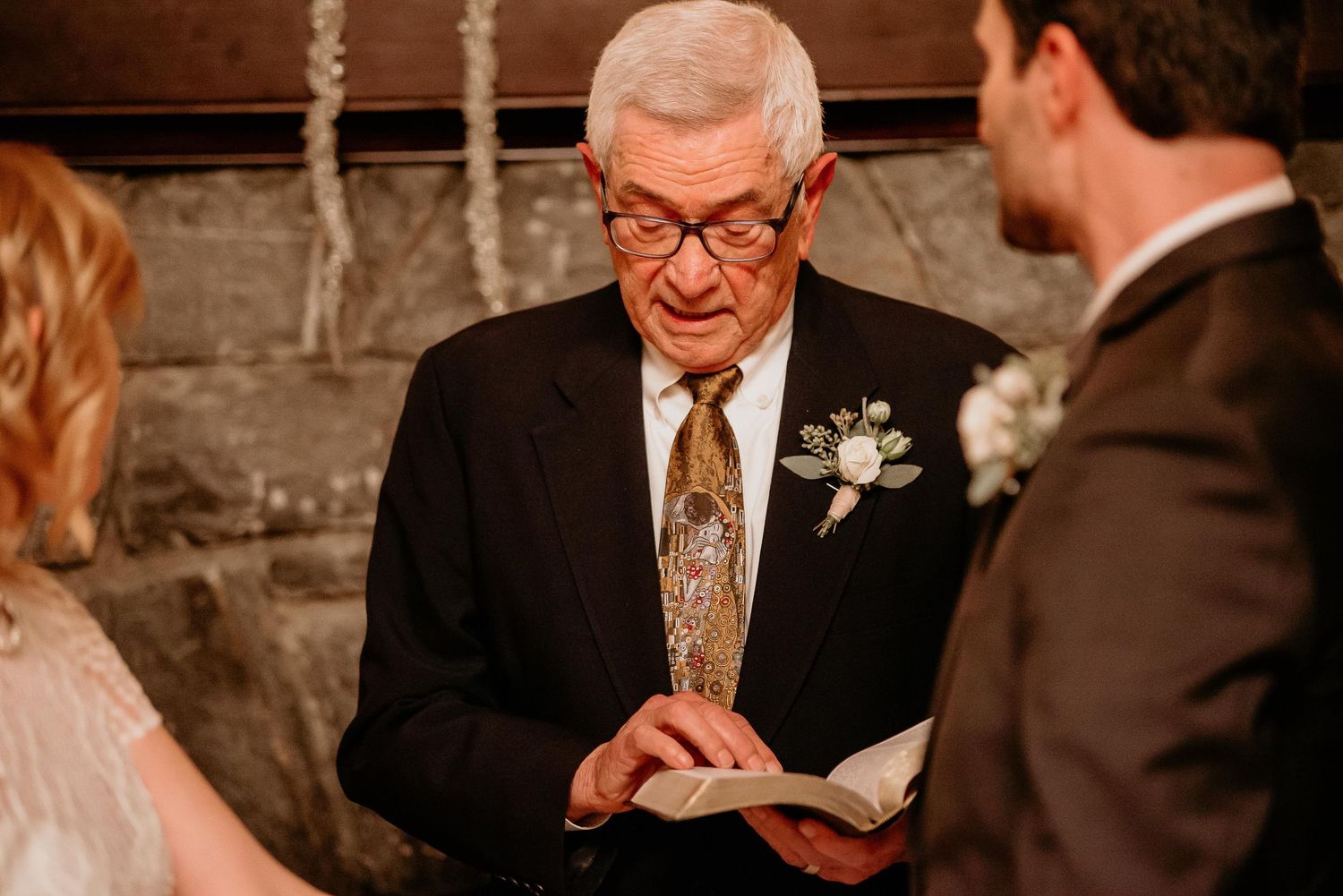Elderly man in dark suit reads from book during wedding ceremony, wearing boutonniere and gold tie.