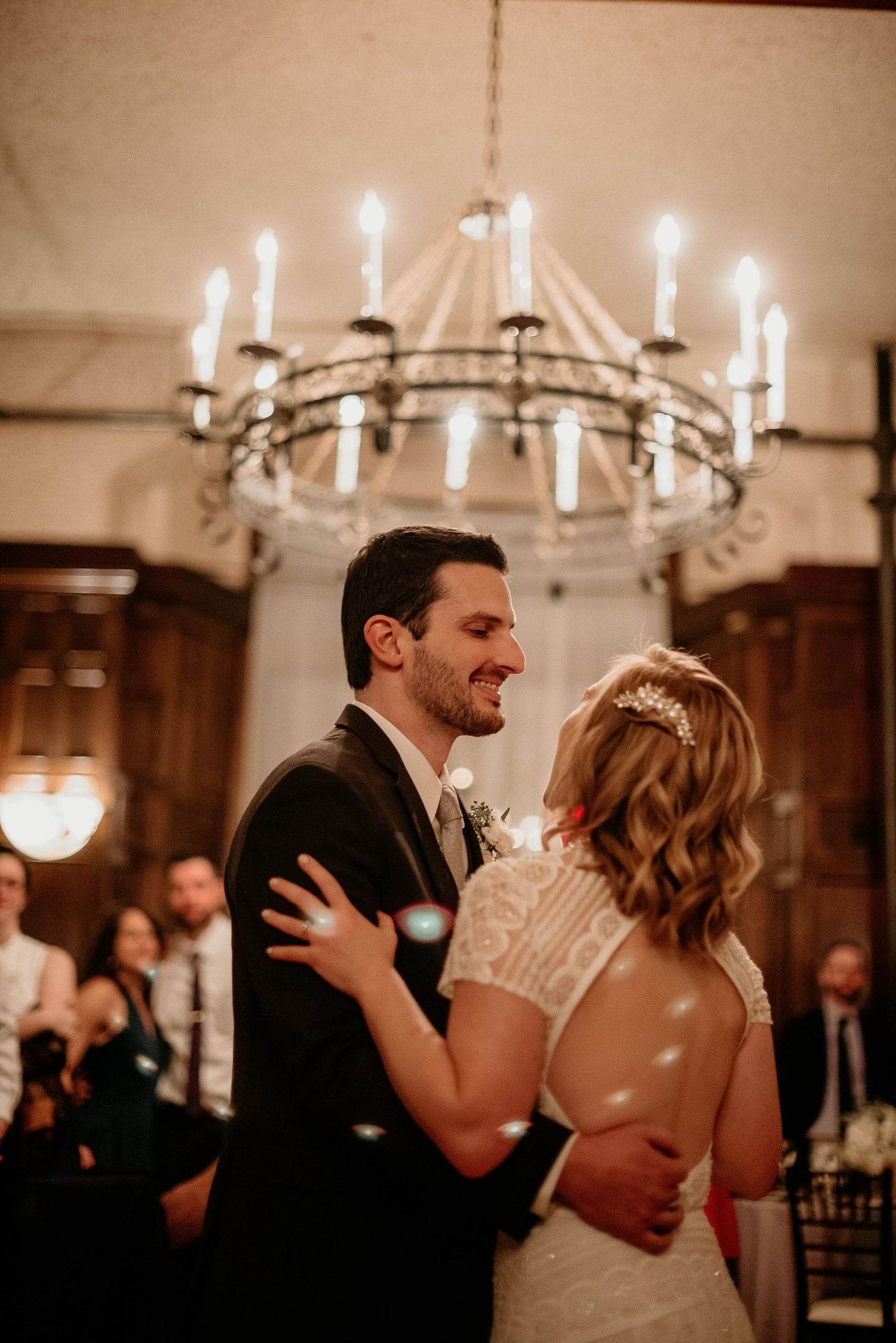 A smiling groom in a dark suit shares his first dance with his bride under an elegant candlelit chandelier at their wedding reception.