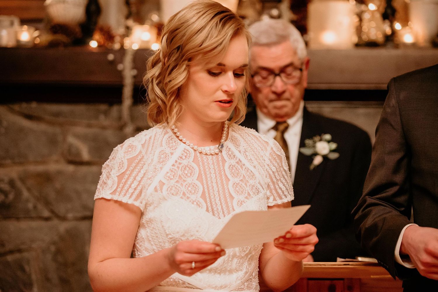 Bride in white lace dress reads vows from paper at wedding ceremony with candles and stone fireplace.