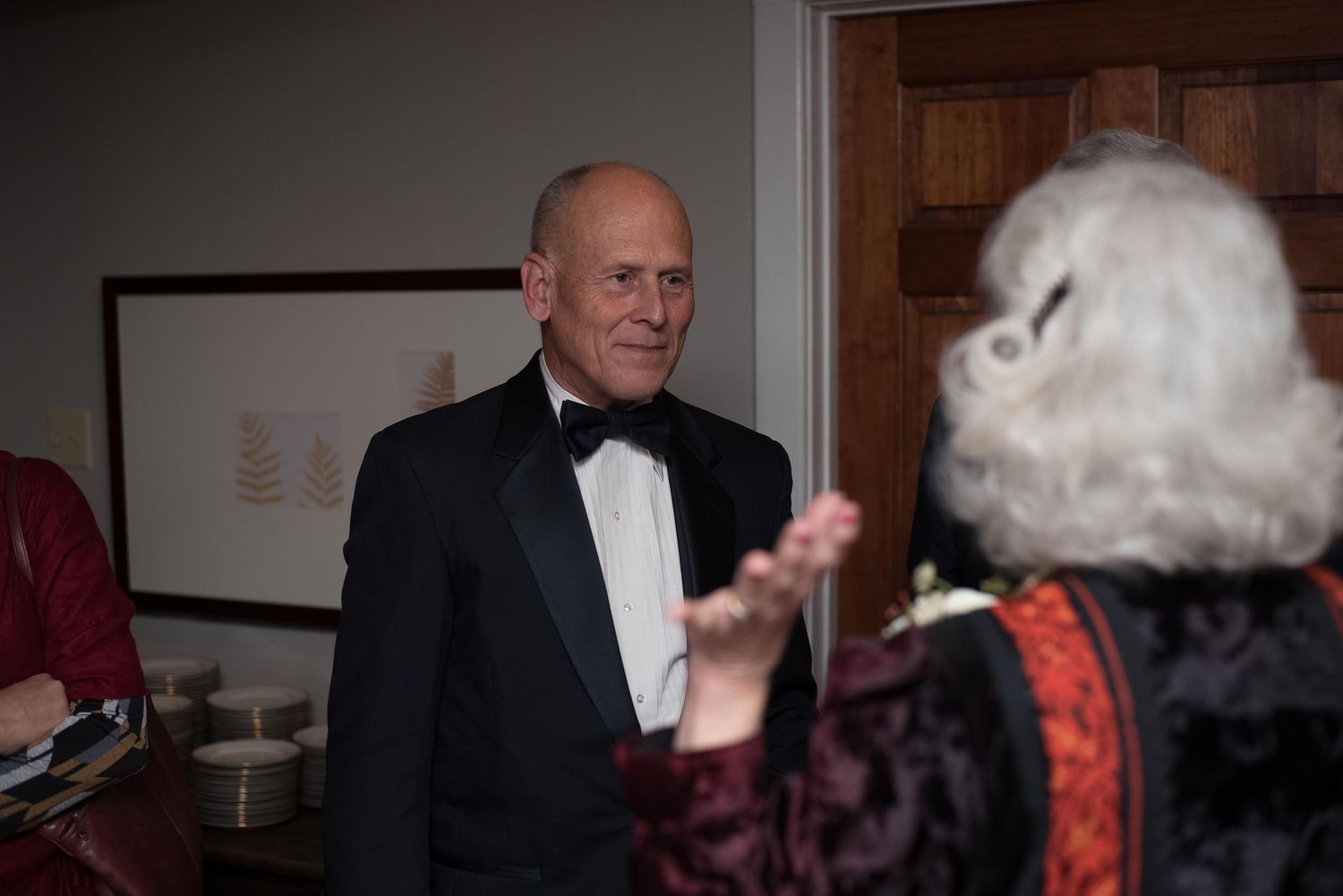 Man in black tuxedo with bow tie conversing with elderly woman in red dress at formal indoor event.