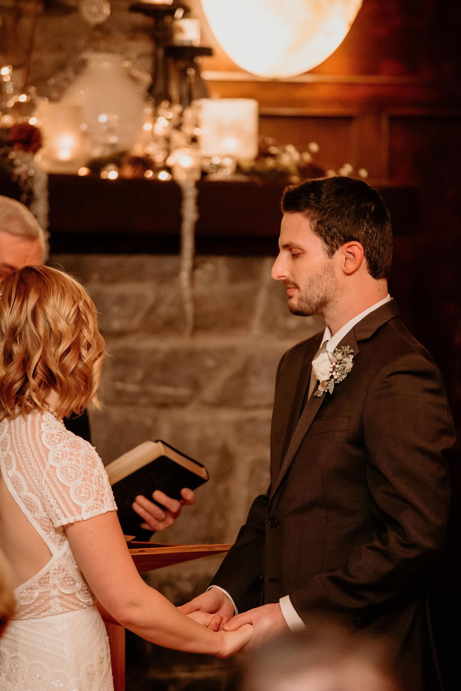 Groom in dark suit holds bride's hands during wedding ceremony by a stone fireplace with warm string lights.