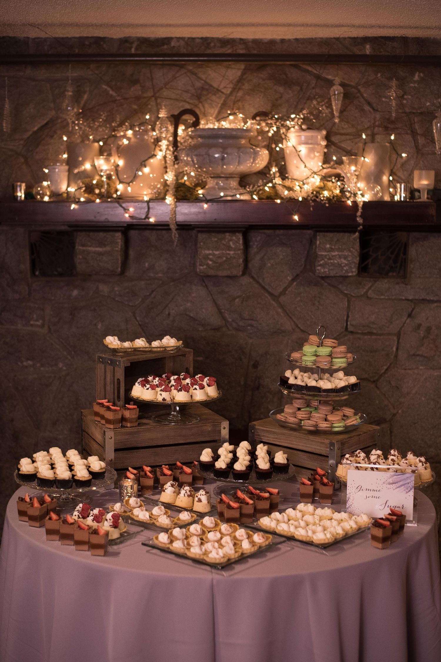 Elegant wedding dessert table with assorted mini treats displayed on wooden crates near a stone fireplace with fairy lights.