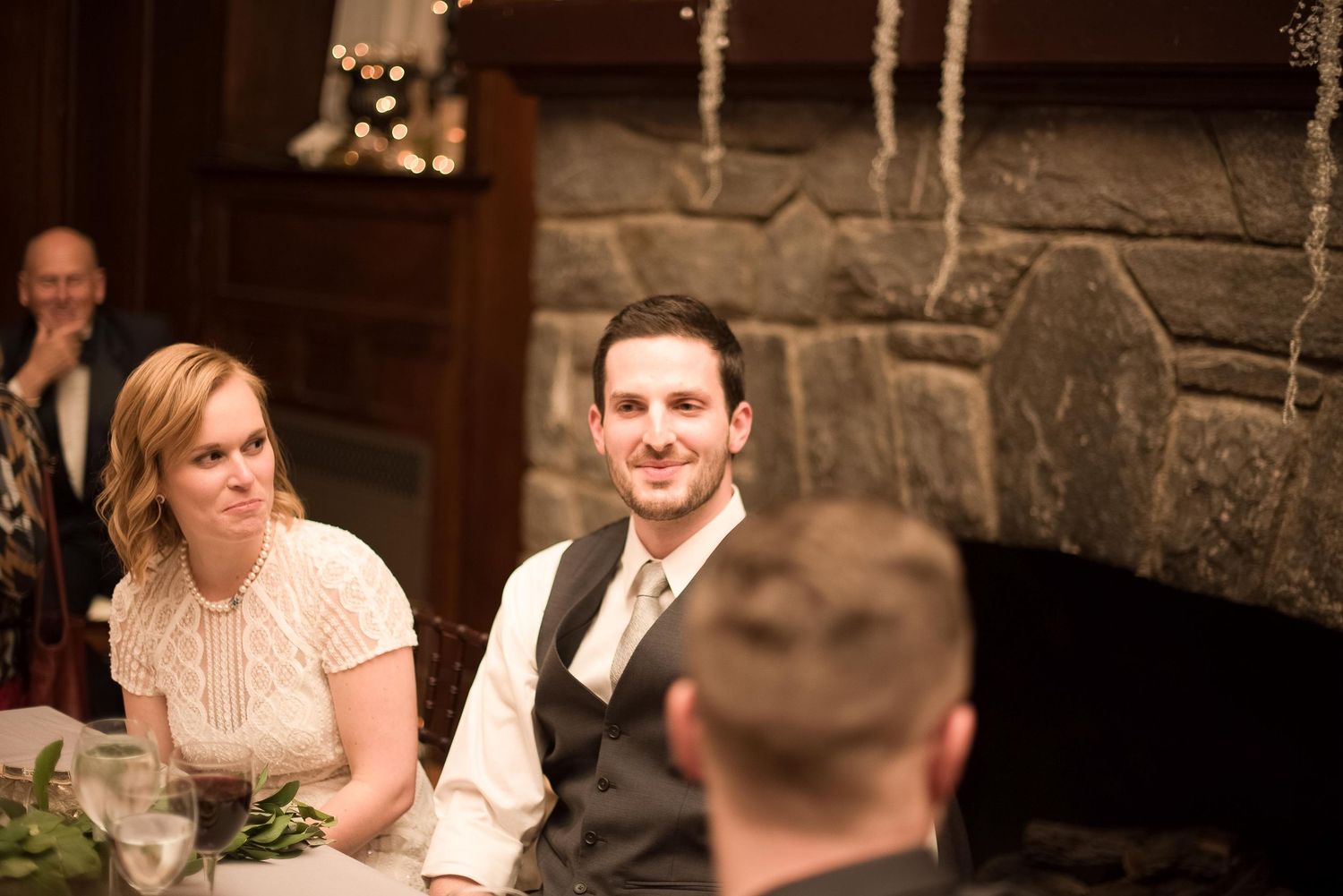 Bride and groom smiling at wedding reception table near stone fireplace.