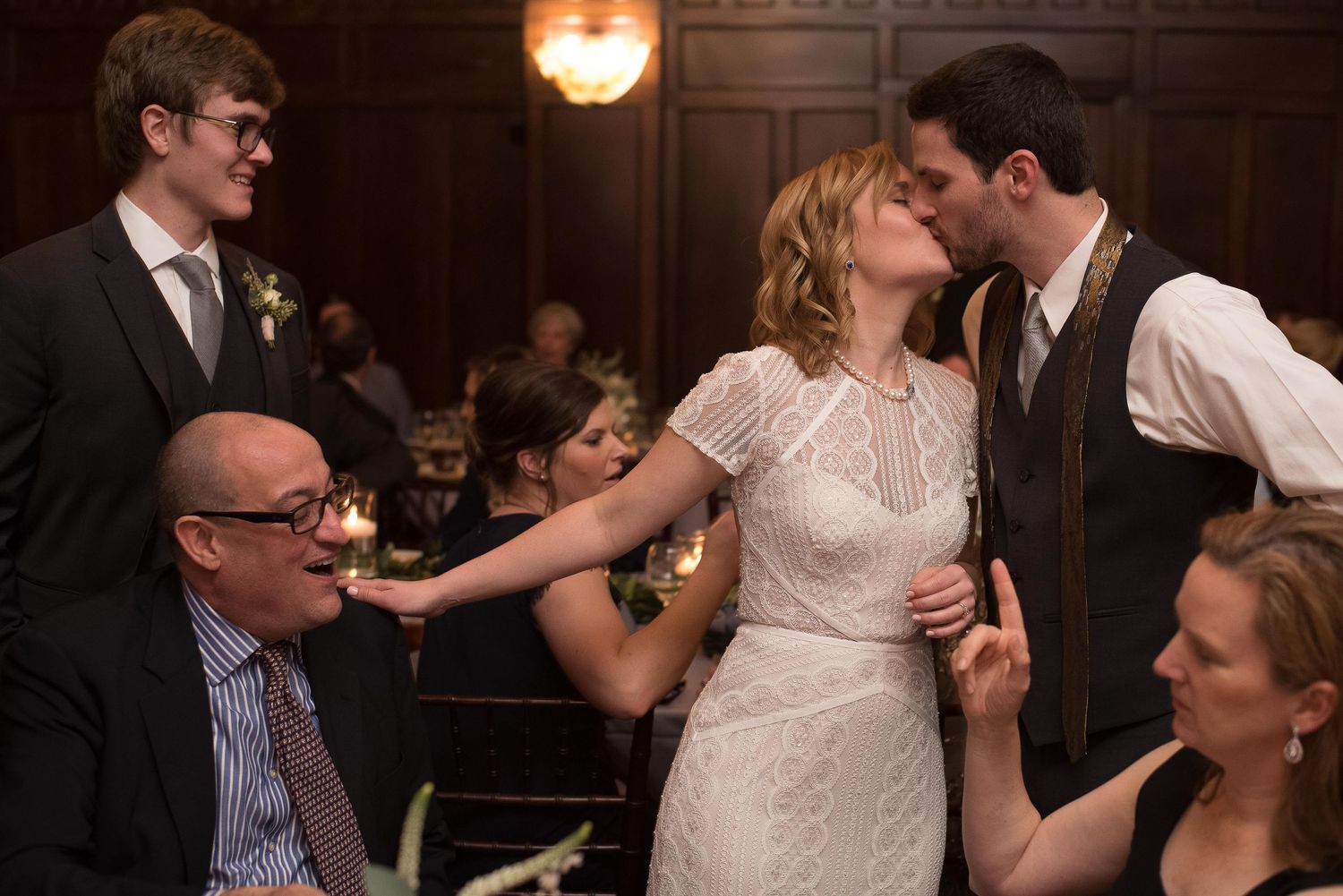Bride in white lace dress shares a kiss with groom in vest while wedding guests celebrate around them.
