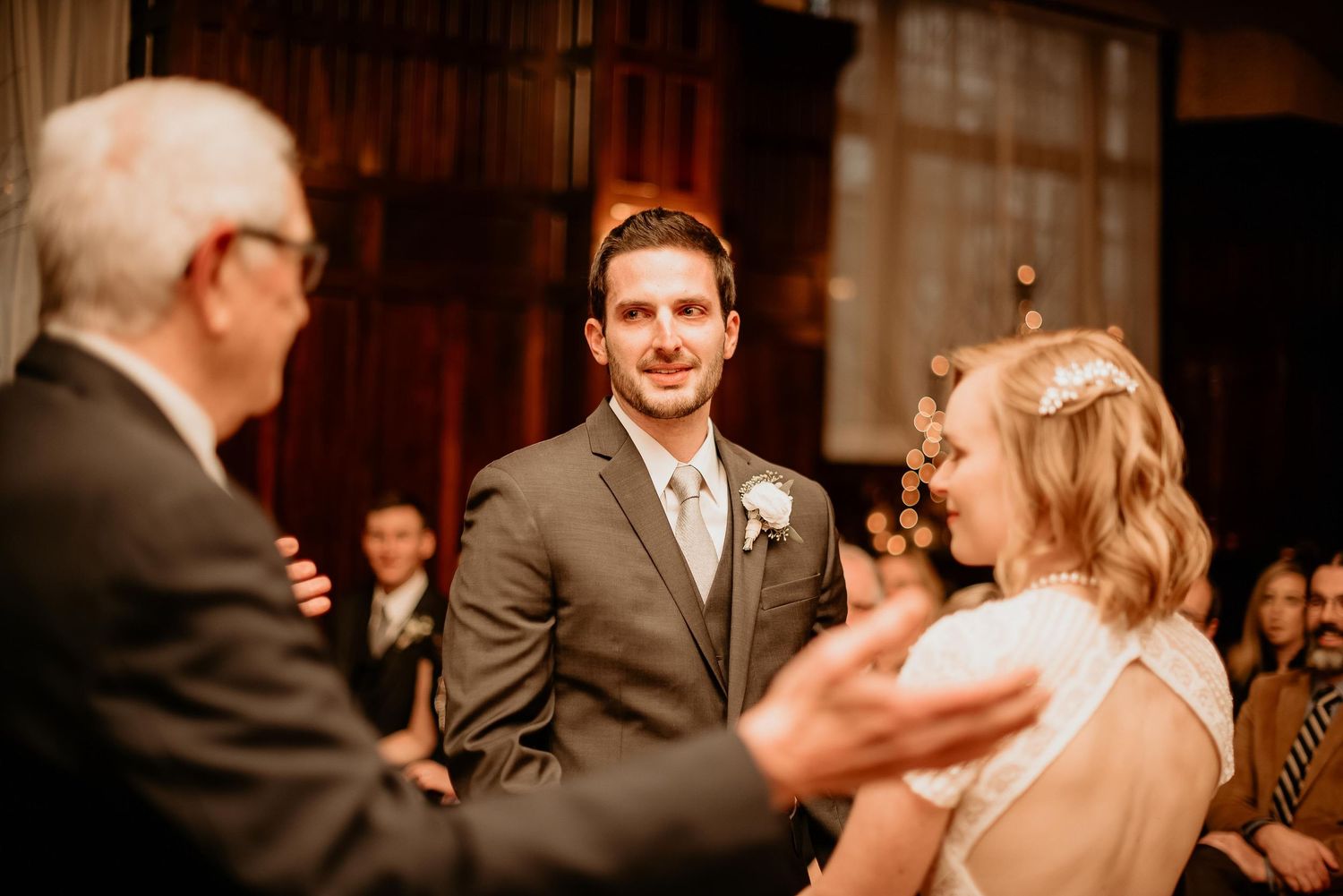 Groom smiling warmly at bride during indoor wedding ceremony with guests seated in background.