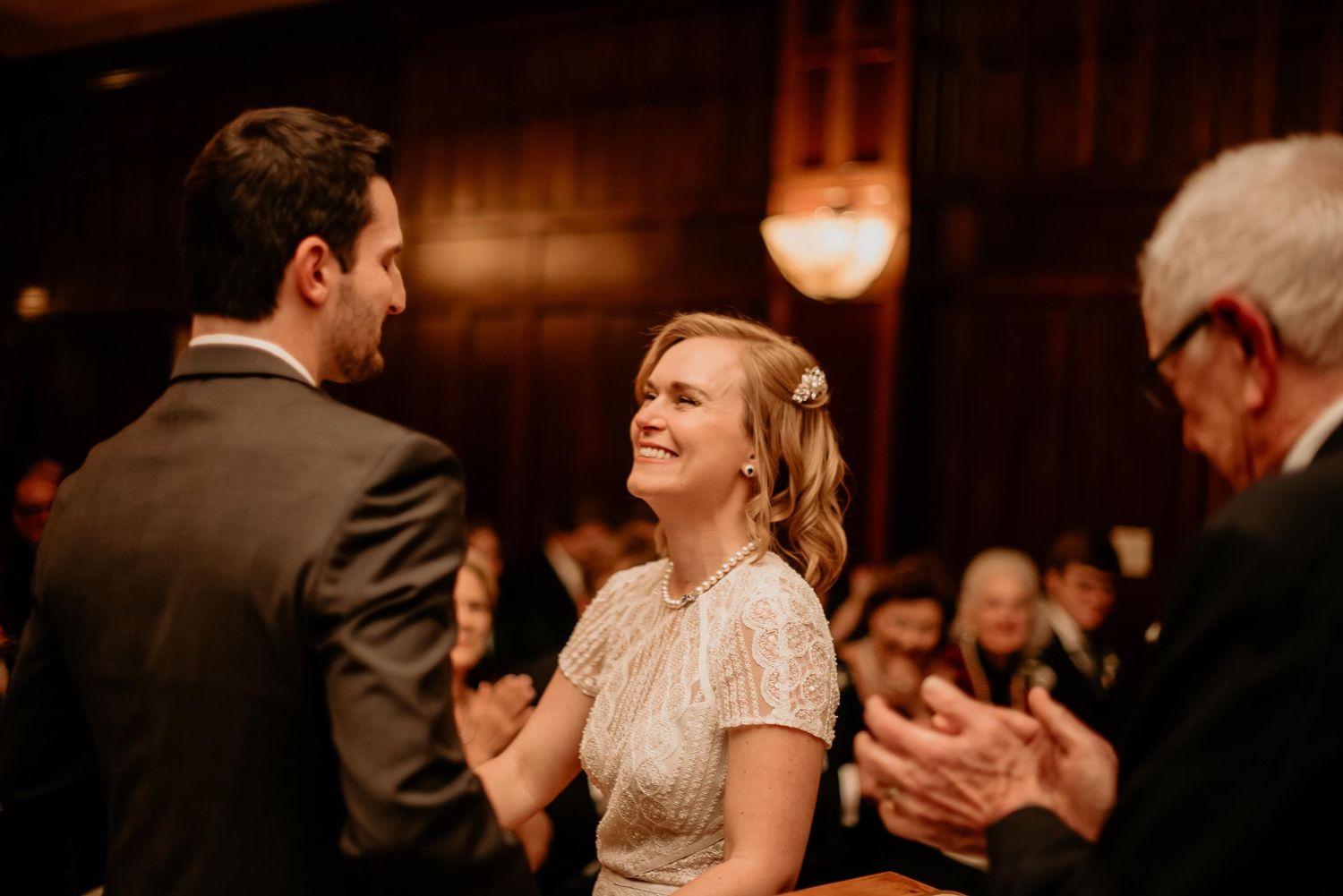 Bride smiling joyfully at groom during wedding reception in elegant wood-paneled venue with guests applauding.