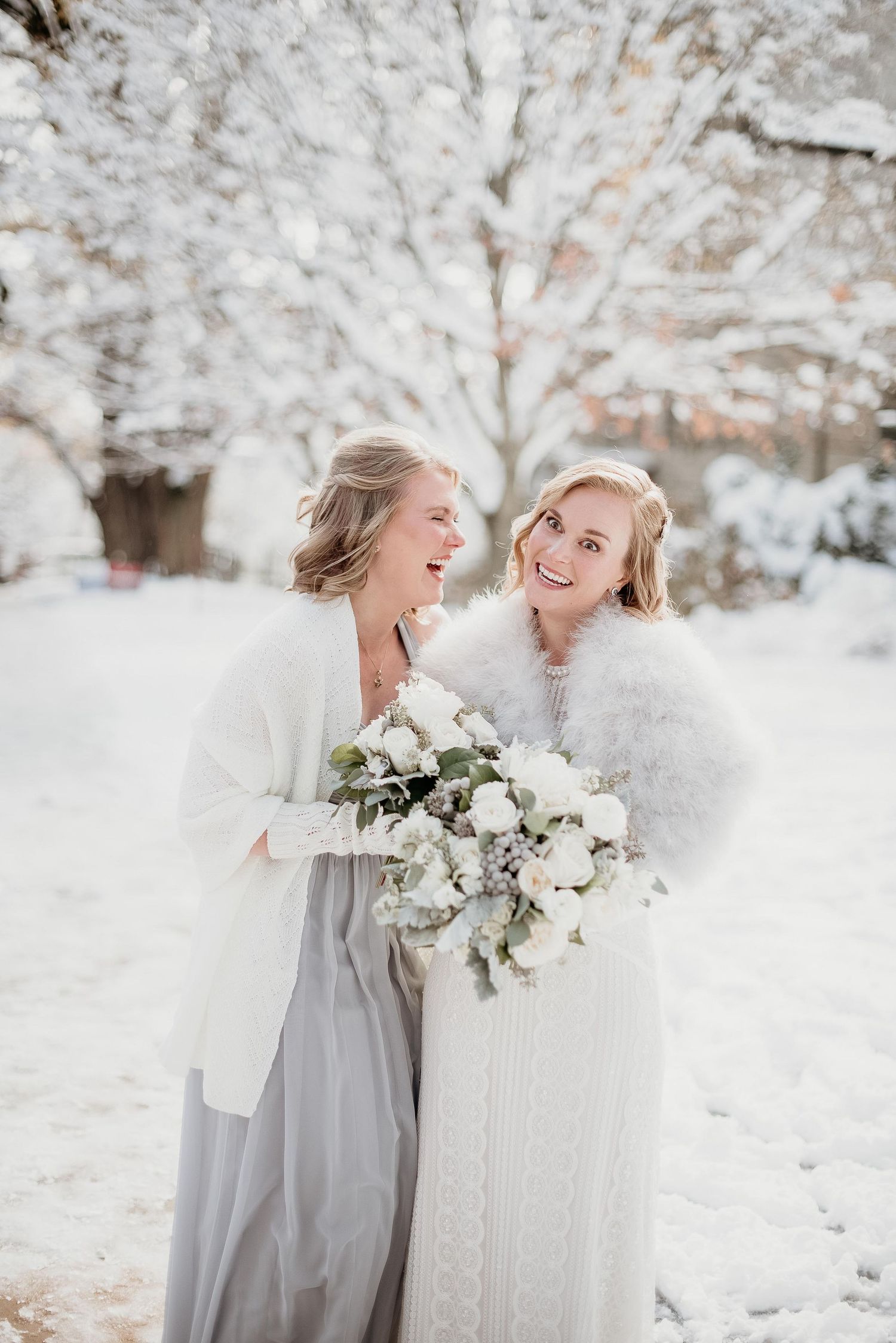 Two brides laughing in snowy winter wonderland, wearing white fur wraps and holding elegant white floral bouquet.