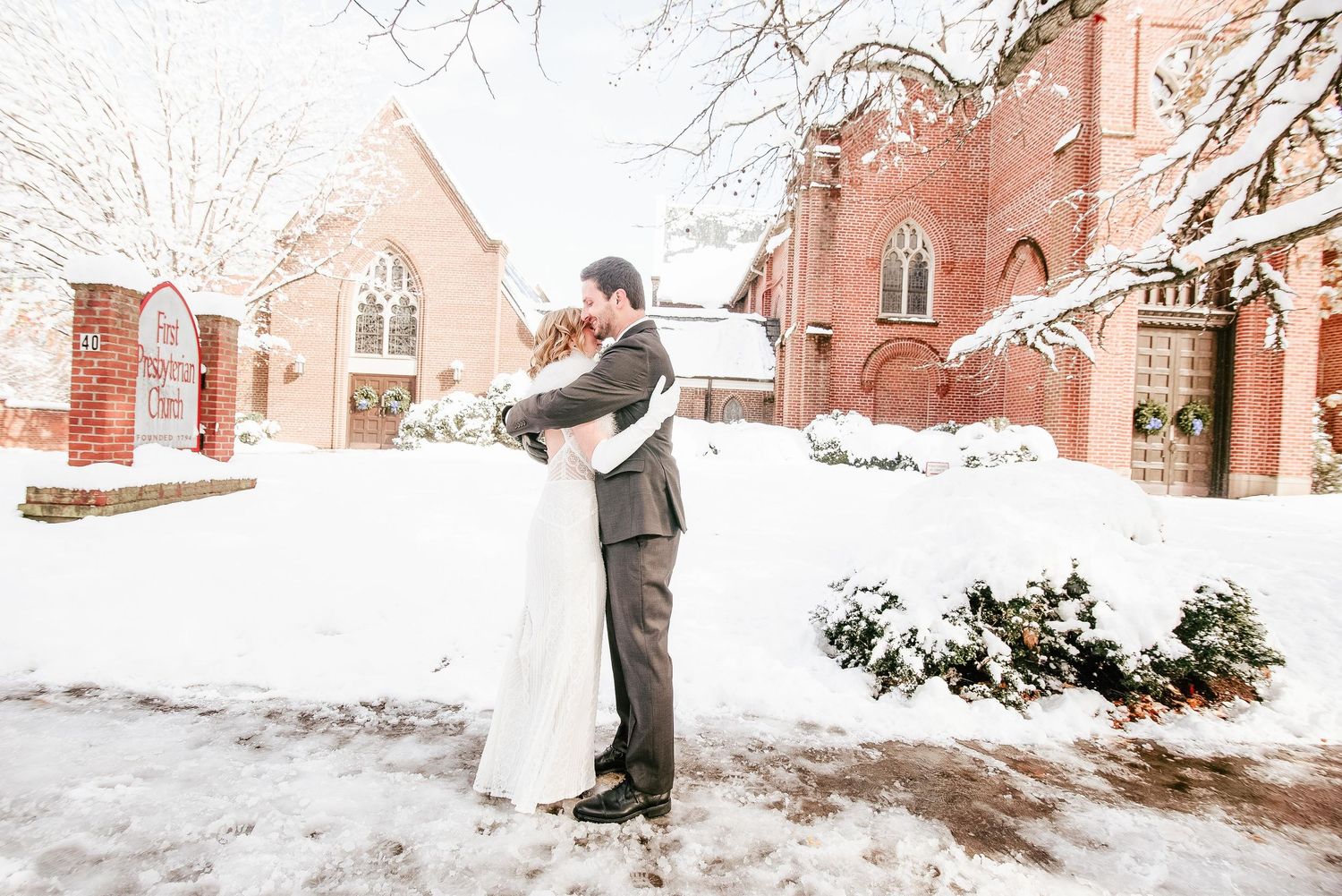 Bride and groom embrace in snowy winter wedding outside red brick church.