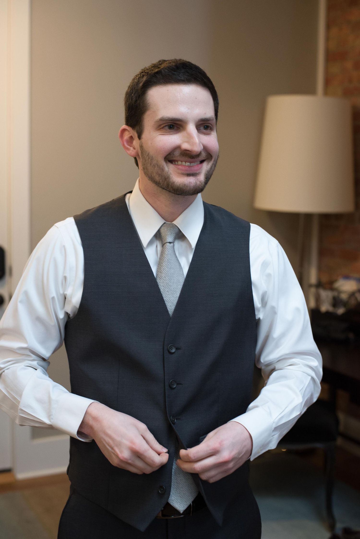 Smiling groom in dark vest and silver tie buttoning his vest while getting ready for wedding.