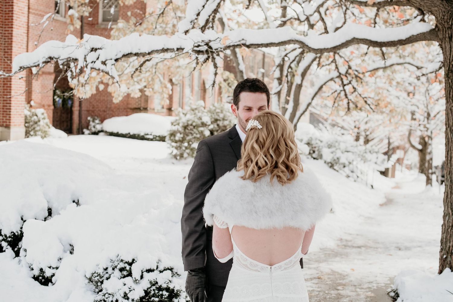 Bride in backless lace dress with fur capelet and groom sharing romantic moment in snowy winter setting.