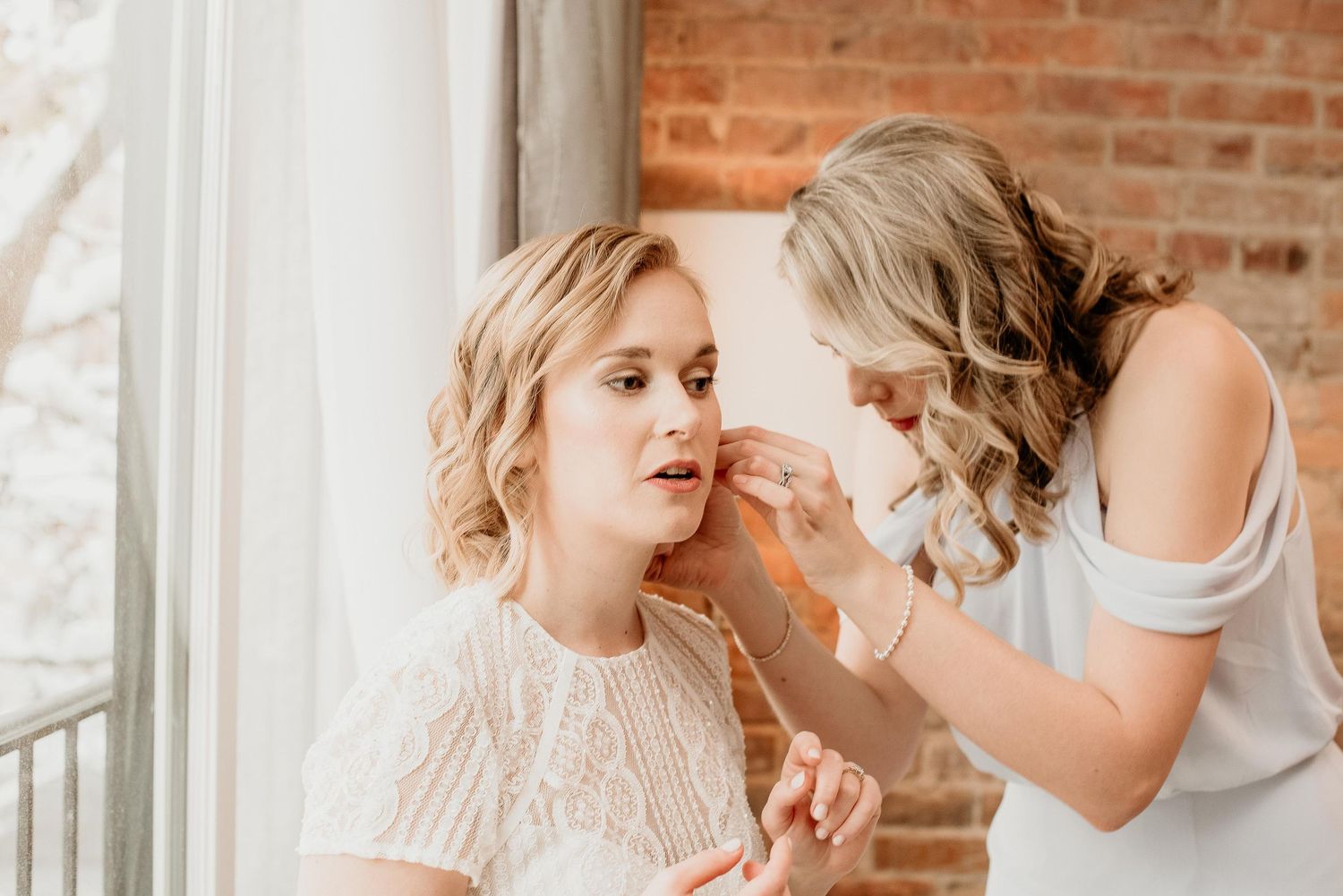 Bridesmaid helping bride put on earrings near a window with brick wall backdrop on wedding day.