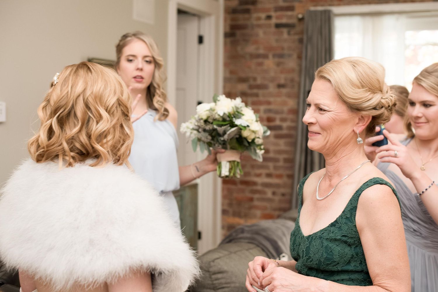 Bride in white fur shawl holding bouquet with bridesmaids and mother in elegant dresses at wedding preparation.