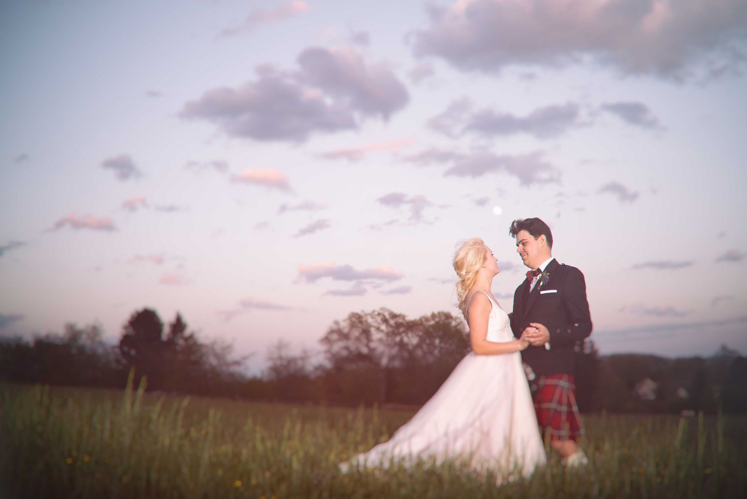 Bride and groom share a romantic moment in a green field at sunset, man wearing a traditional Scottish kilt.