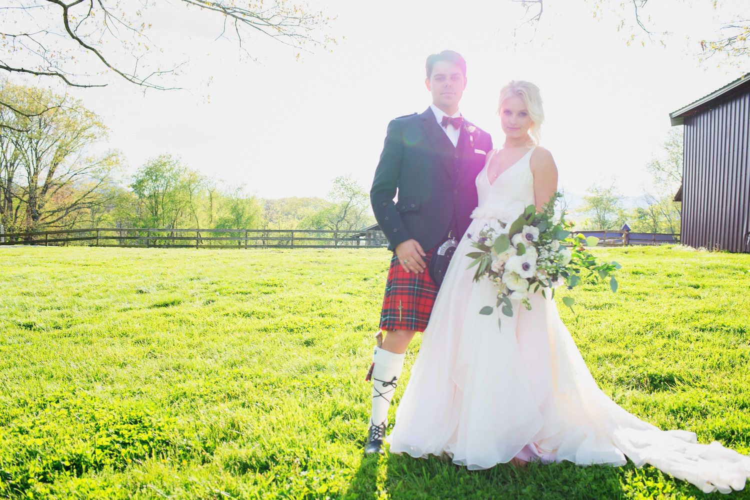 Bride and groom in Scottish kilt posing on lush green lawn at outdoor wedding venue on sunny day.