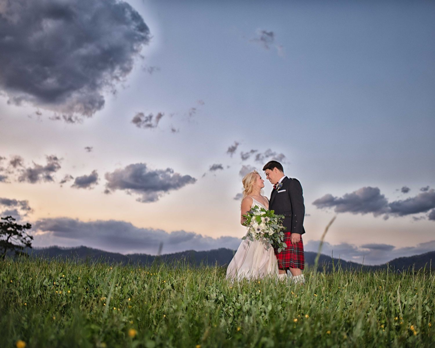 Bride and groom share a romantic moment in a green meadow at sunset, groom wearing a red tartan kilt.