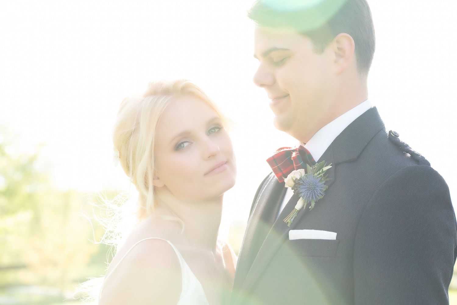 A bride and groom share a romantic moment outdoors, bathed in soft sunlight, with the groom in a dark suit and red bow tie.