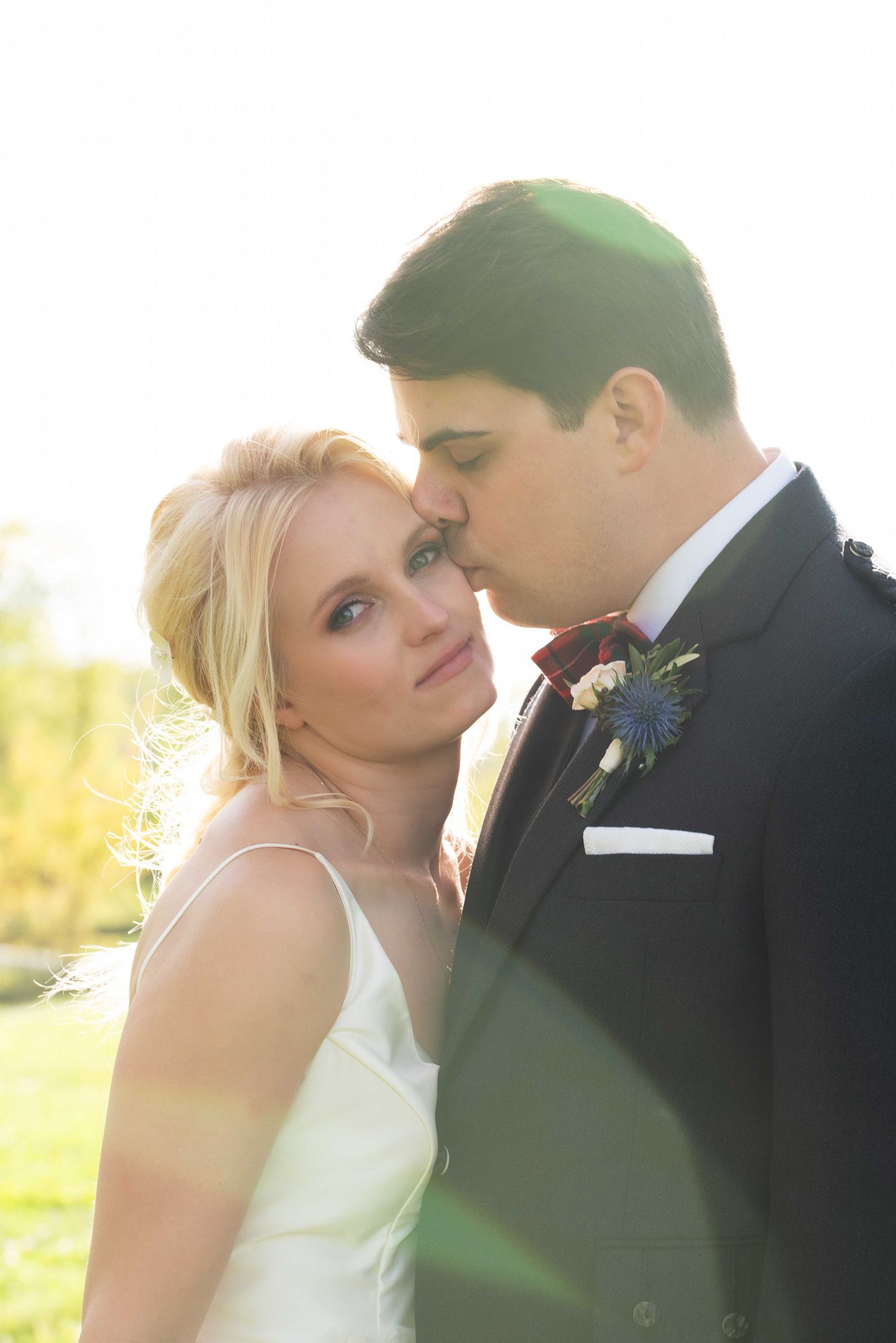 Groom in black suit kissing bride on cheek outdoors in golden sunlight, bride in white dress smiling at camera.