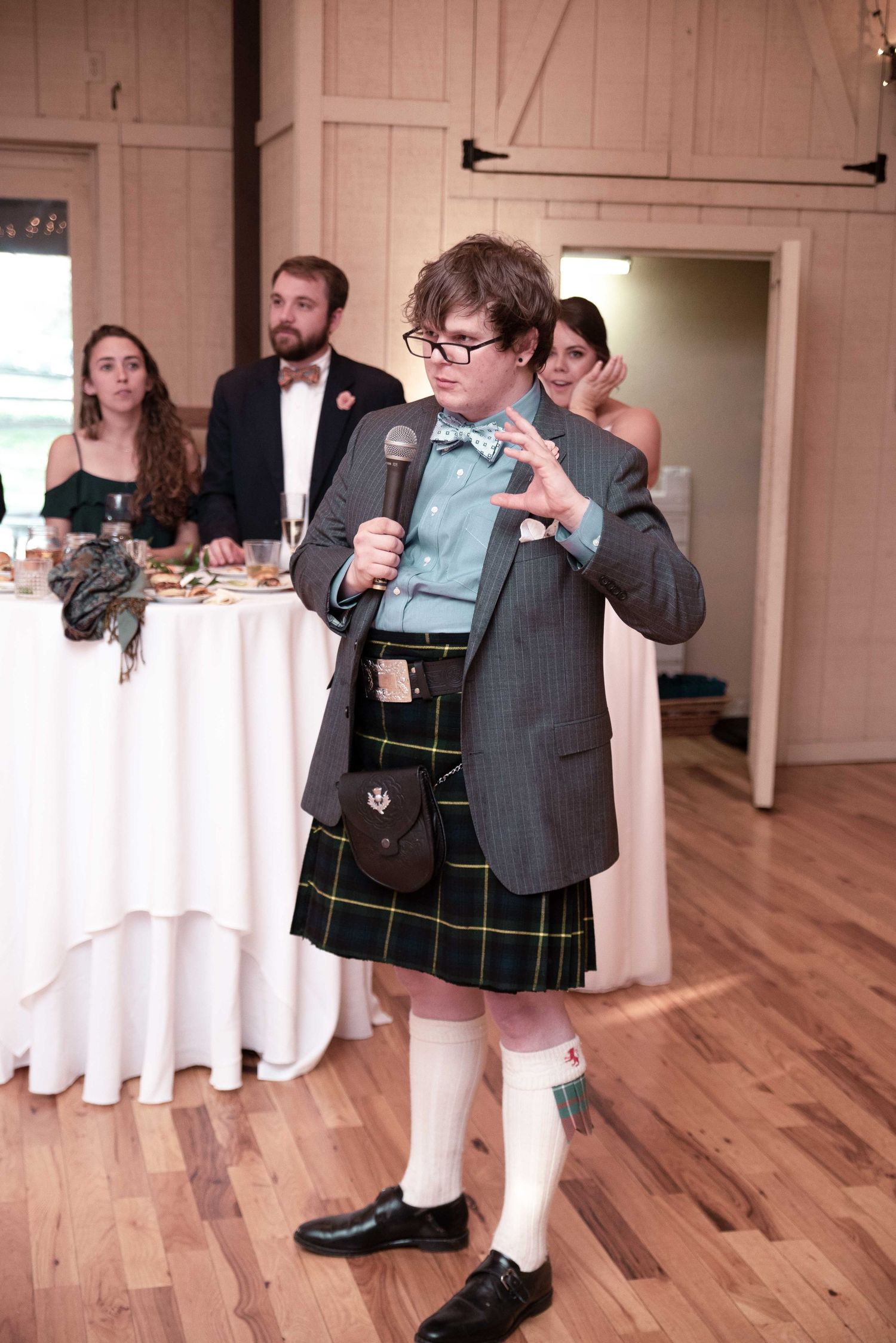 Person in kilt, blazer, and bow tie giving a speech at a wedding reception while holding a microphone.