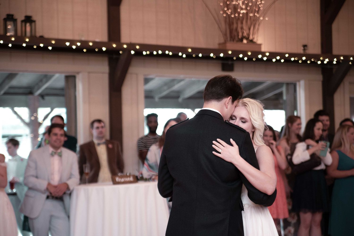 Bride and groom share their first dance at a rustic barn wedding reception with string lights and guests watching.
