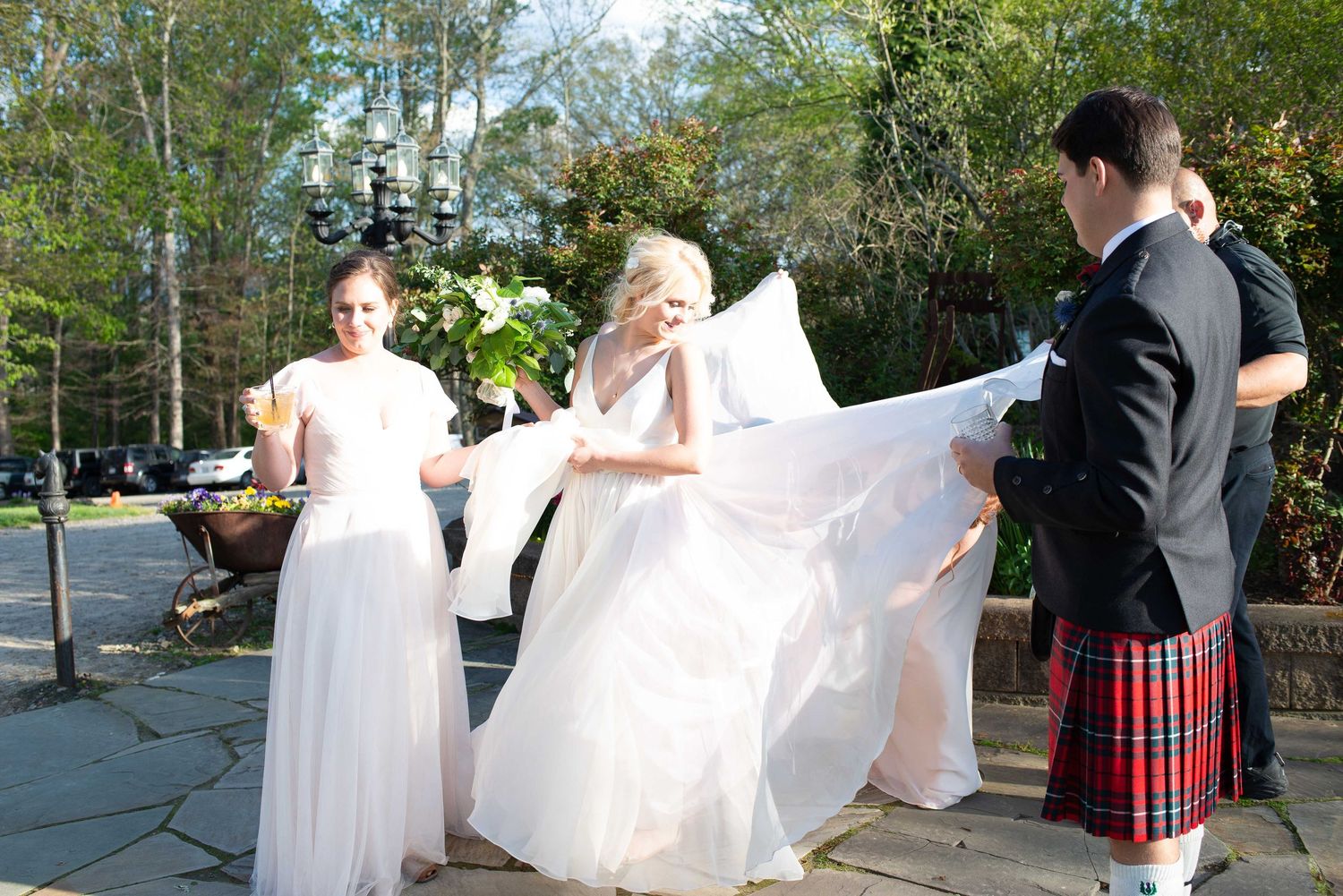 Bride in white gown with bridesmaids and groom in red tartan kilt adjusting wedding dress outdoors at a garden venue.