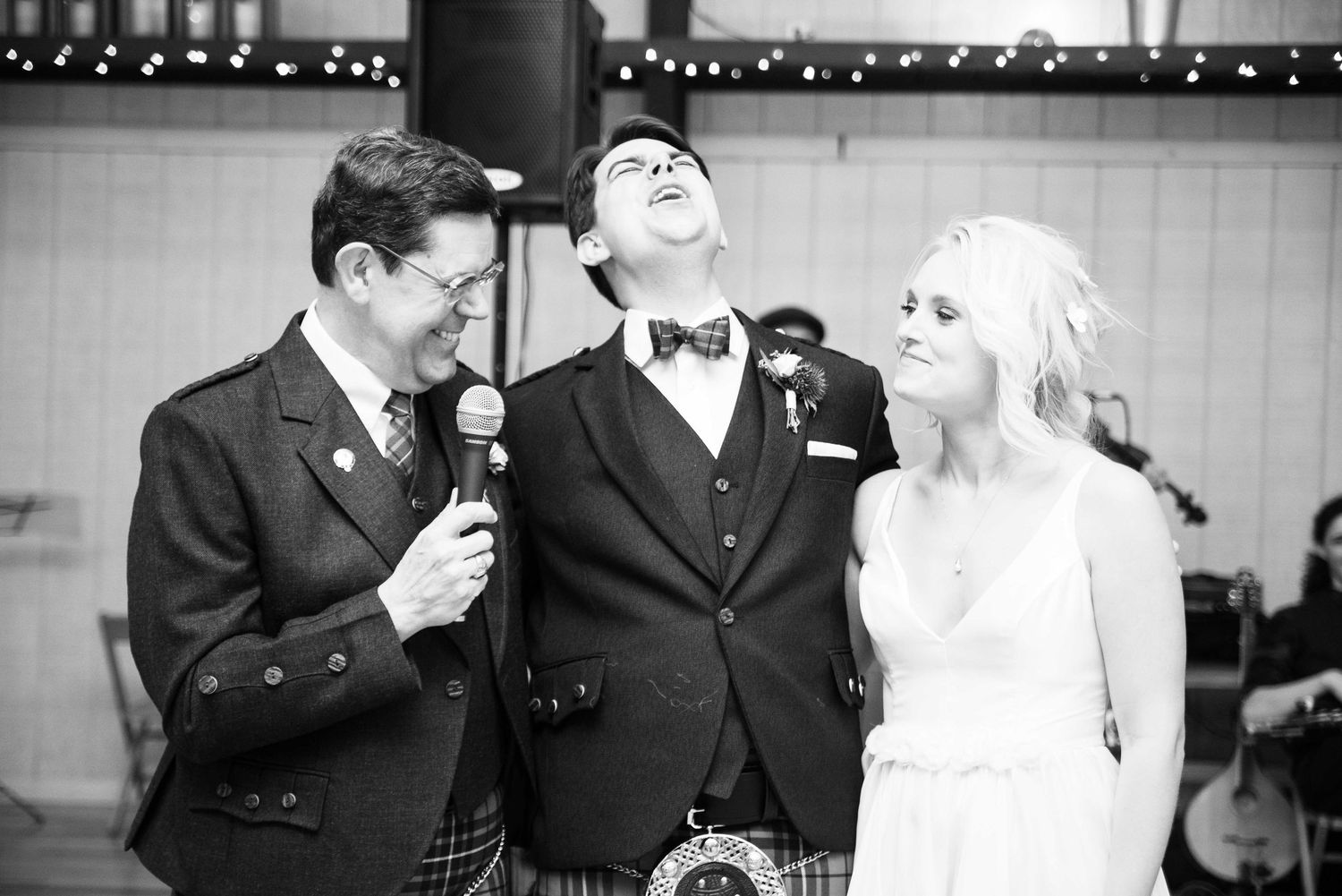 Black and white photo of a bride and groom laughing with a man in a kilt holding a microphone at a wedding reception.