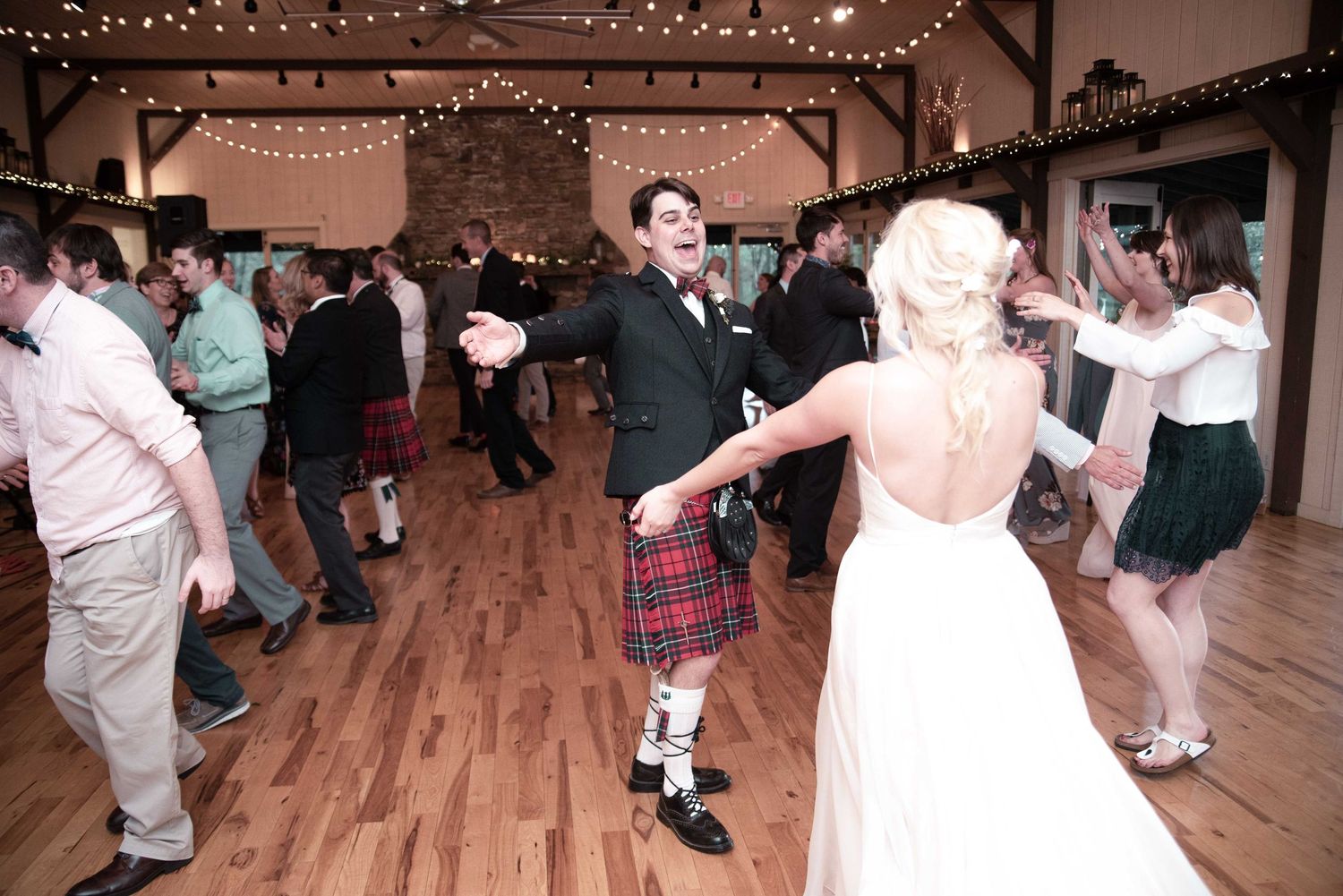 Groom in red tartan kilt dances with bride in backless white gown at rustic barn wedding reception.