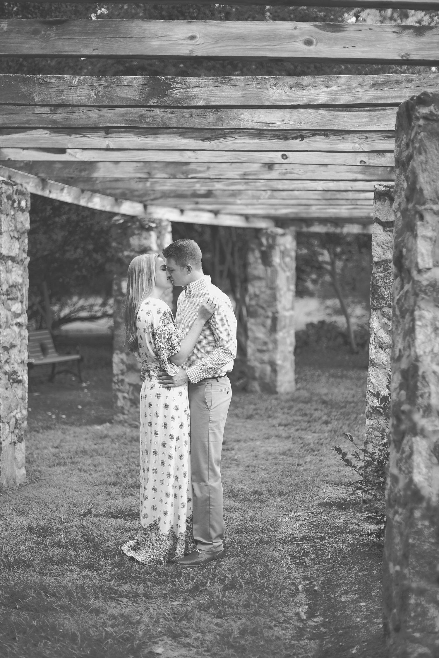 Black and white photo of a couple sharing a romantic moment under a rustic wooden pergola with stone pillars.