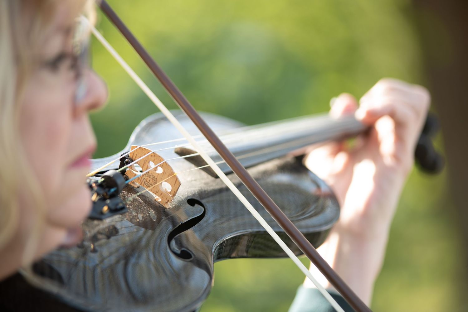 Close-up of blonde woman playing violin at outdoor wedding with blurred green trees in background.