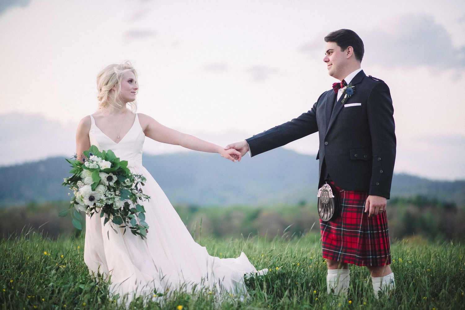 Bride in white gown holding hands with groom in red tartan kilt in a green meadow with mountain backdrop.