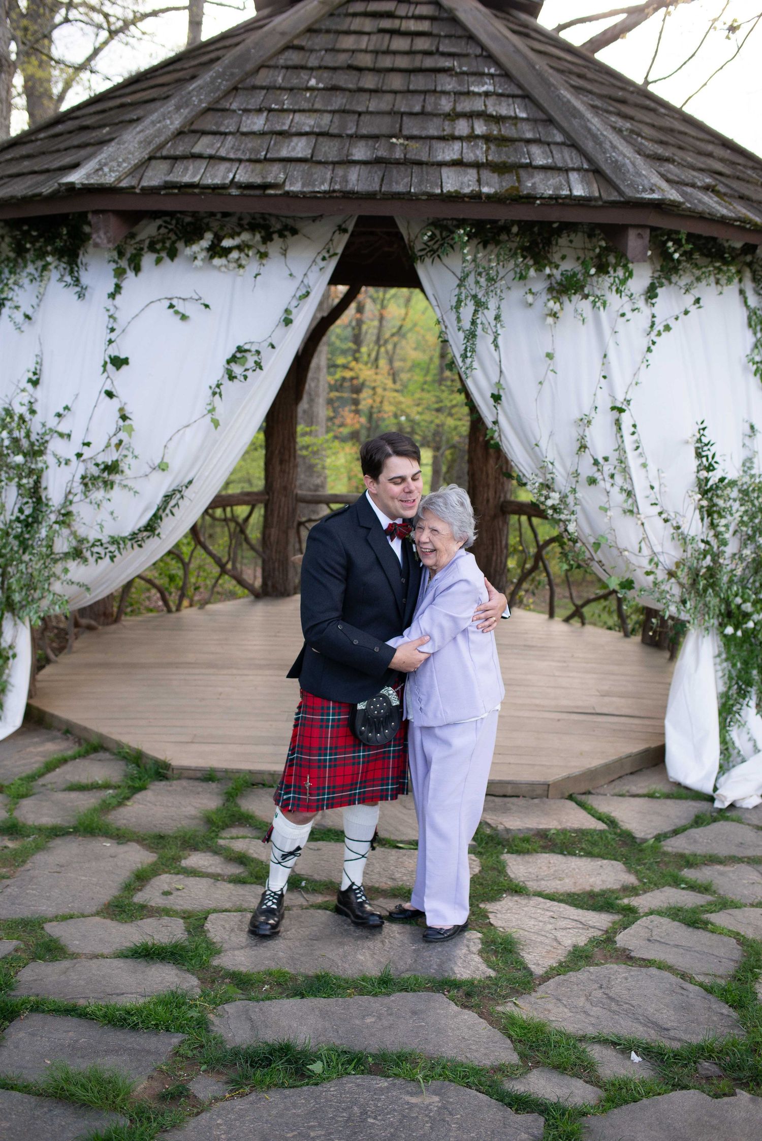 Man in Scottish kilt and tuxedo embraces elderly woman under decorated gazebo with white draping and greenery.