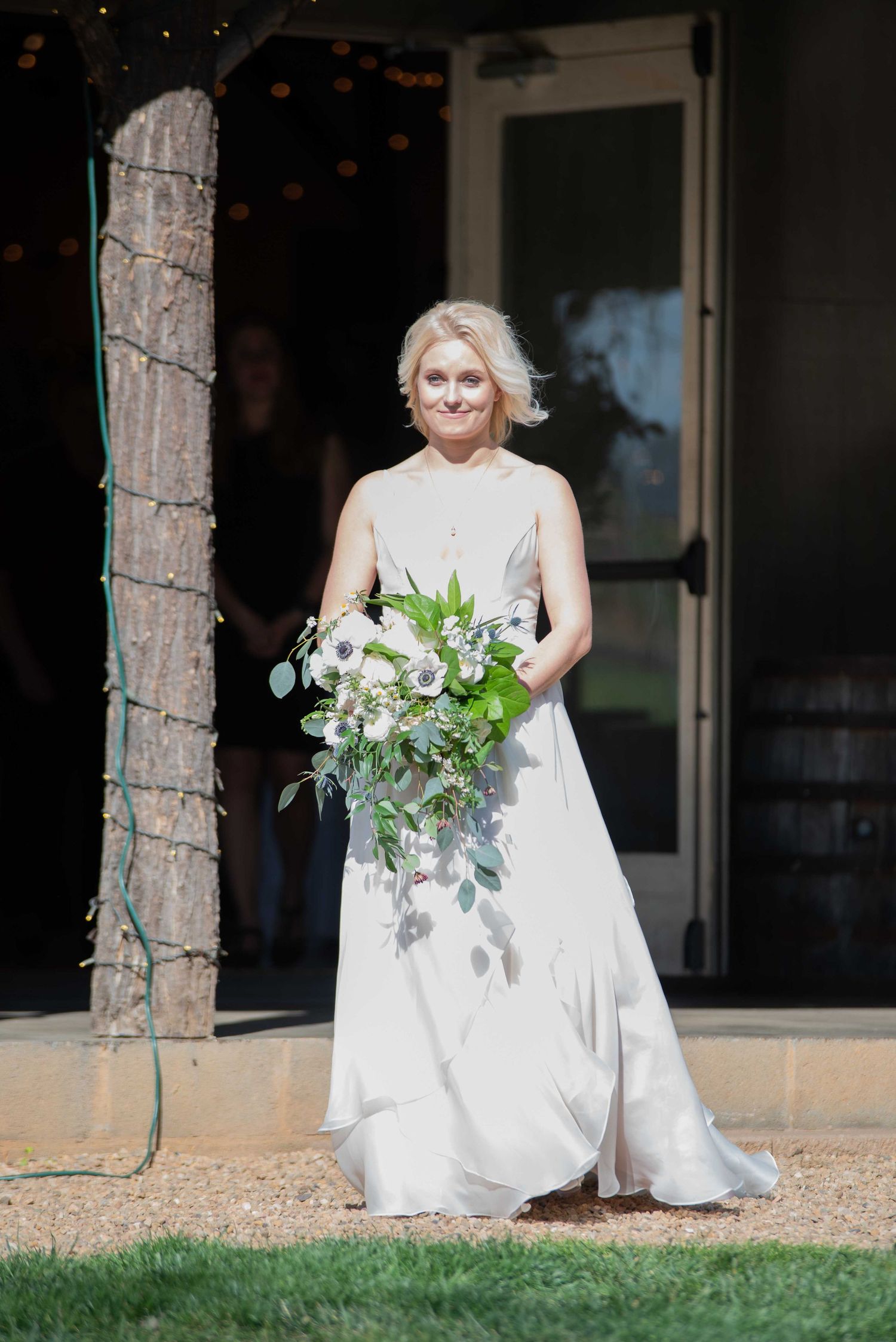 Bridesmaid in elegant gray gown holding white and green bouquet walking outdoors at wedding venue.