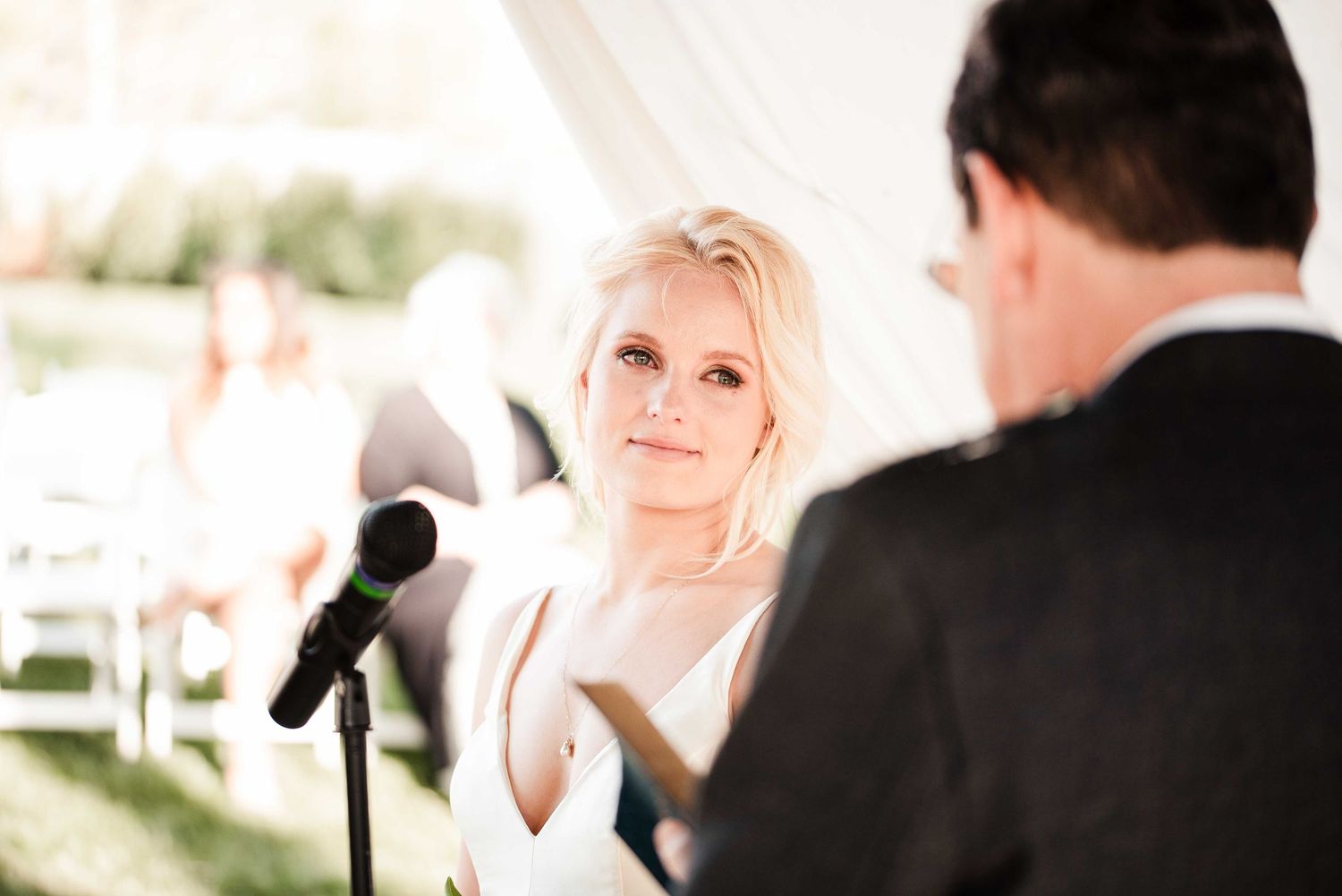 Bride gazing lovingly at groom during outdoor wedding ceremony under white tent with microphone in foreground.