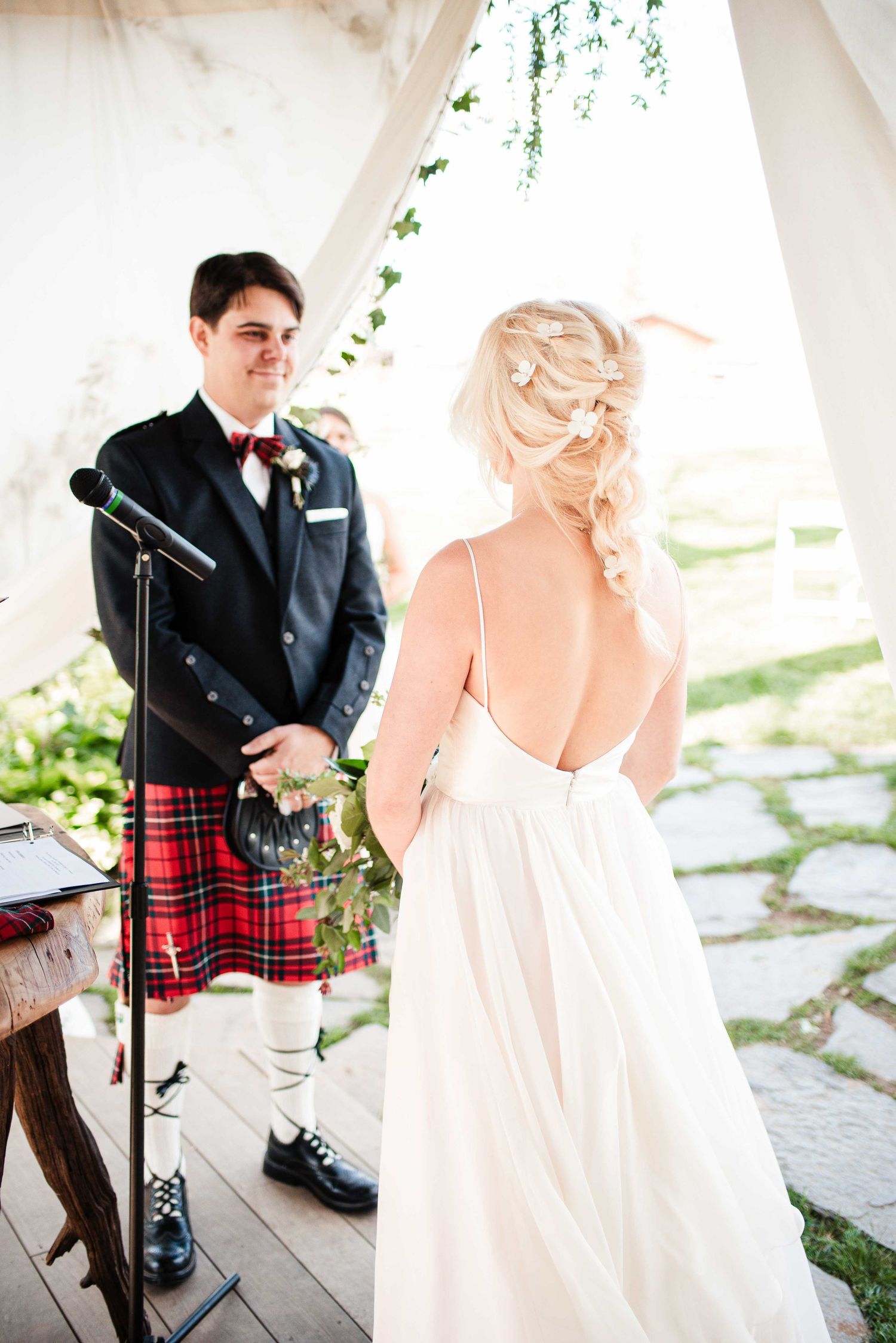 Groom in Scottish kilt and bow tie smiles at blonde bride in backless white gown during outdoor wedding ceremony.